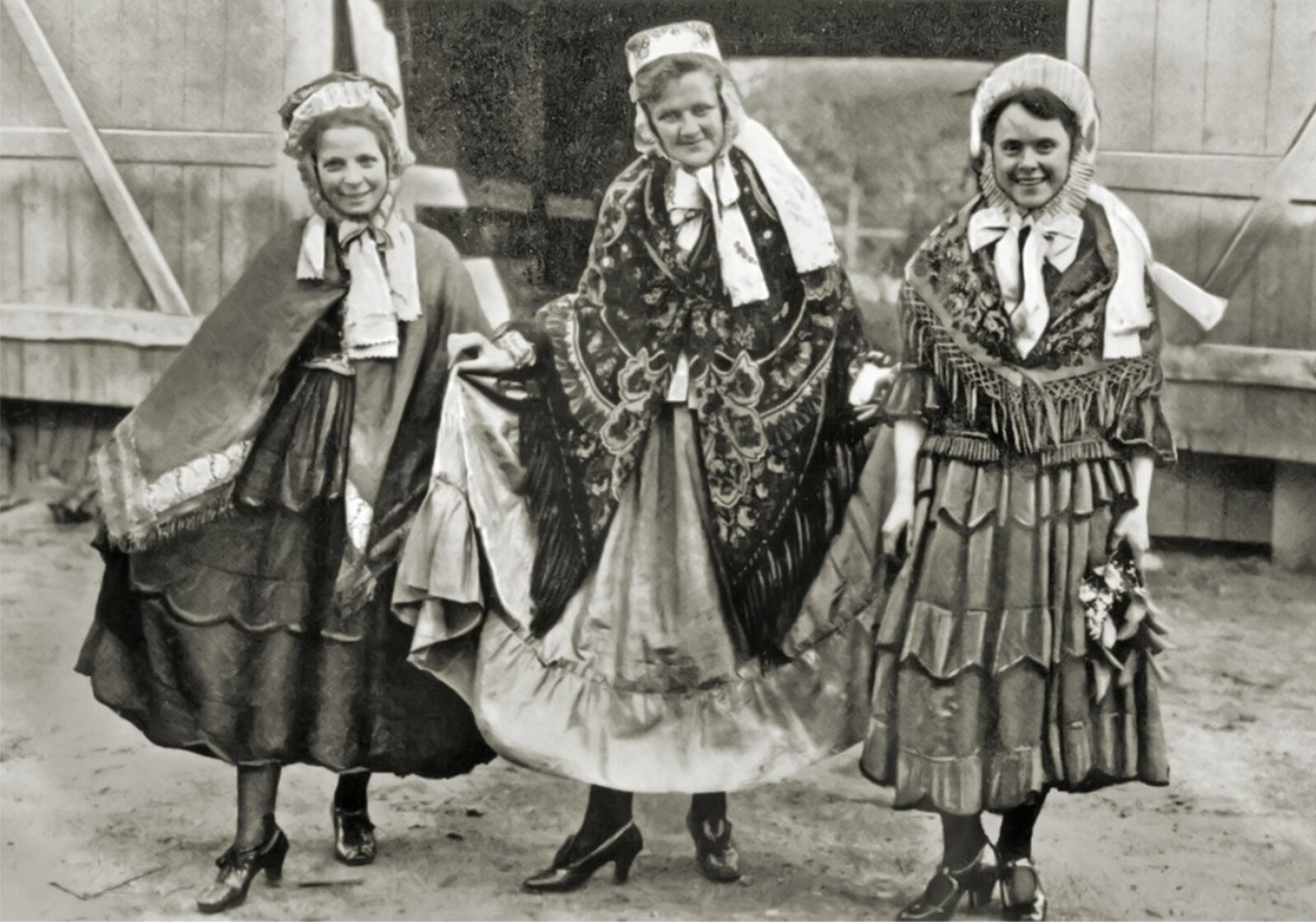 Czarno‑biała fotografia przedstawiająca trzy kobiety w obszernych, bogato zdobionych sukniach. Na ich ramionach widać duże chusty. Kobiety noszą również nakrycia głowy przypominające czapki, wiązane pod szyją.