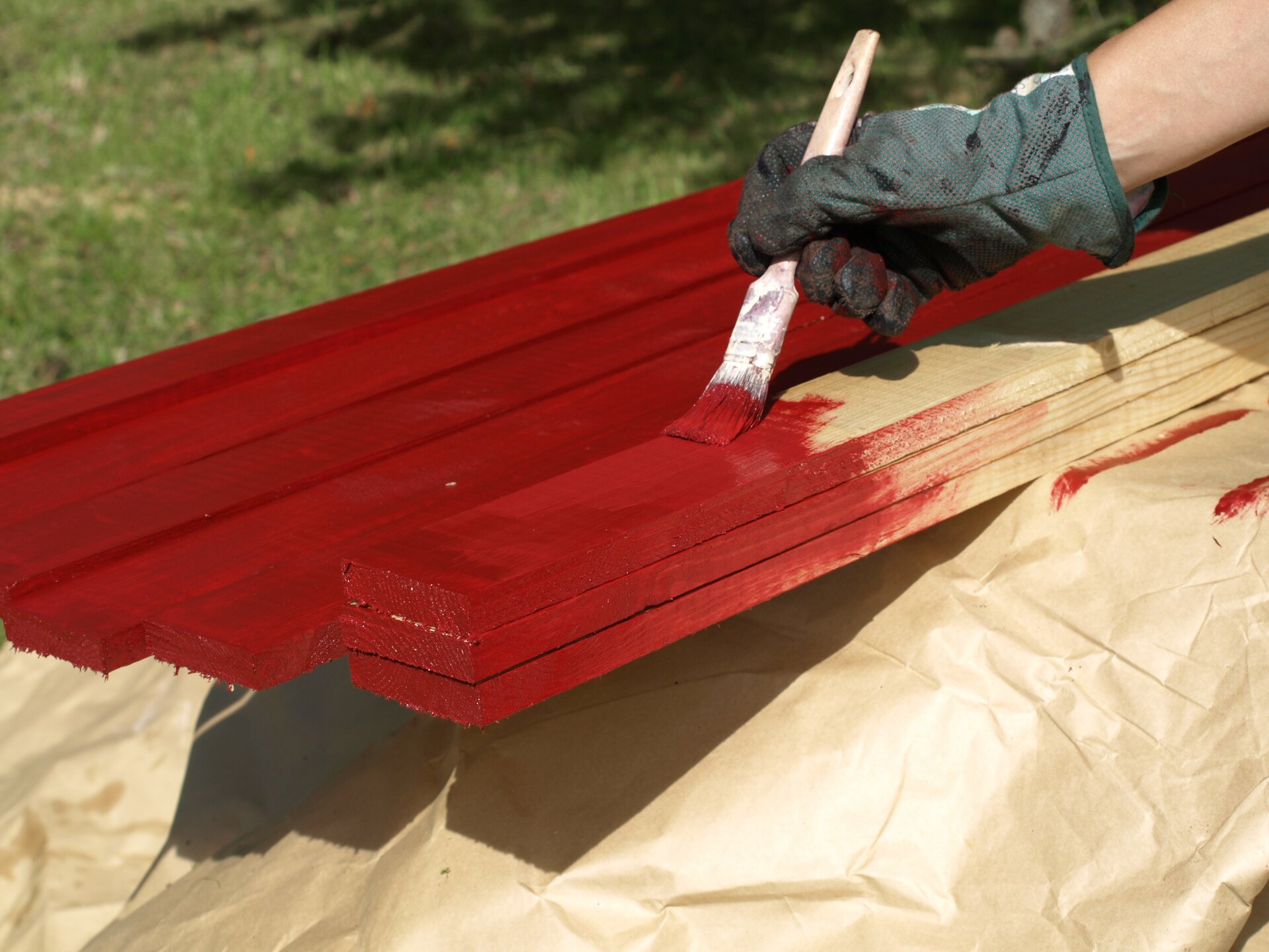 Ilustracja przedstawia deski malowane czerwoną farbą. Nad deskami znajduje się ręka w rękawiczce, która pędzlem maluje deski. W tle znajduje się trawa oraz cień drzewa.