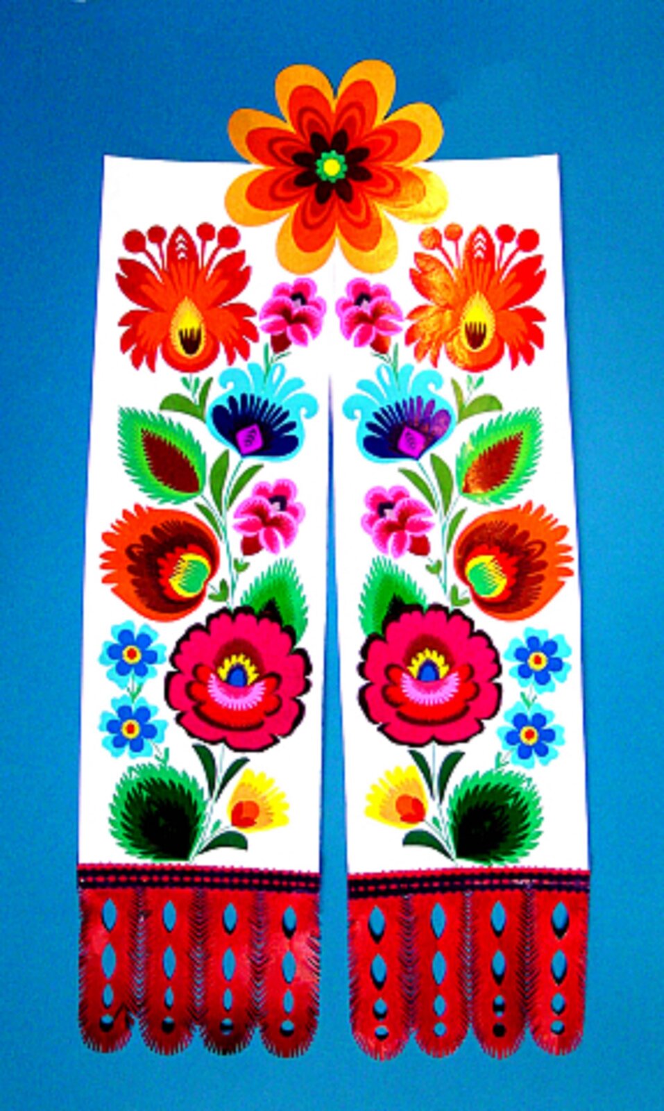 Ilustracja przedstawia łowicką tasiemkę. Są to dwie takie same części, zakończone są czerwonymi wypustkami. Na obu znajdują się kwiaty w różnych kolorach oraz zielone listki. Wzór ułożony jest pionowo. Dwie taśmy łączy znajdujący się pośrodku górnej części kwiat.