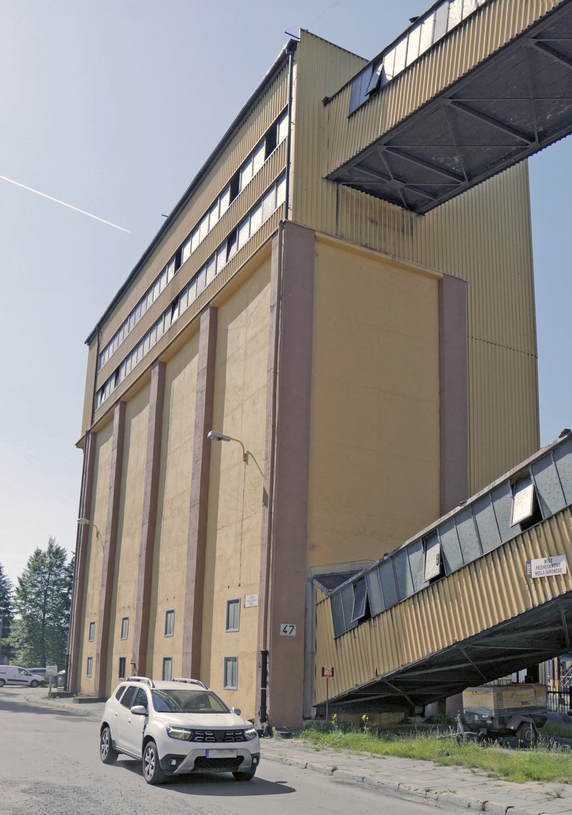 Na zdjęciu widoczny jest teren przemysłowy pokazany dołu. Po lewej stronie zdjęcia widoczny jest duży prostopadłościenny budynek. Do podstawy budynku przyłączony jest łącznik. Łącznik znajduje się pod kątem. Łącznik wykonany jest z żółtej blachy falistej. W górnej części ściany osadzone są okna. Na boku łącznika widoczna jest tablica z napisem: most przenośnikowy węgla surowego. Przy budynku zaparkowany jest samochód.Na terenie placu widać części urządzeń oraz metalową konstrukcję przypominającą ramę przenośnika.