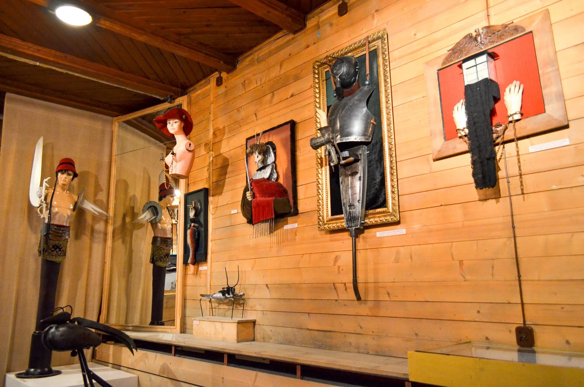 Zdjęcie przedstawia wnętrze galerii artystycznej. Na ścianie wiszą dzieła w ramach, z trójwymiarowymi elementami. Dzieła składają się z różnych materiałów i mają nieregularne kształty. Między innymi jest to zbroja, szalik, plastikowe dłonie. W kącie pomieszczenia stoją dwa popiersia manekinów - kobieta i mężczyzna, ubrani w nakrycia głowy i inne elementy.