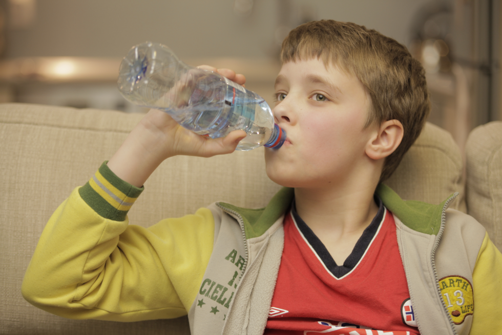 Fotografia przedstawiająca chłopca pijącego wodę z butelki. Chłopiec ubrany jest w sportowy strój kurtkę oraz T-shirt. Chłopiec siedzi na kanapie, butelkę trzyma w prawej dłoni. Chłopiec w wieku około dziesięciu lat.