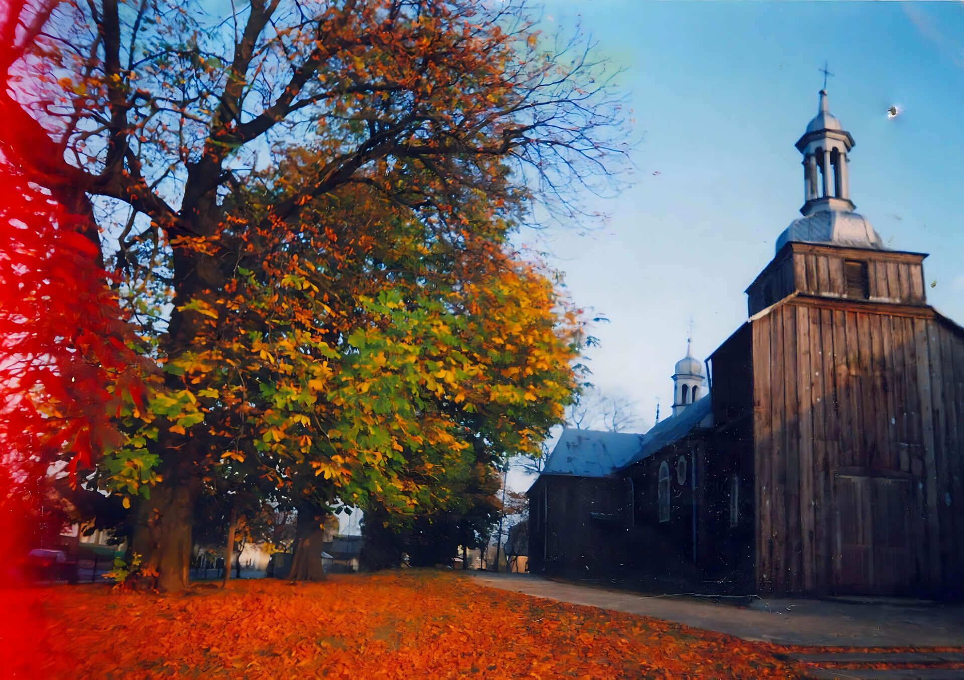 Zdjęcie przedstawia drewniany kościół ze skośnym, srebrnym dachem. Na dachu znajdują się dwie wieżyczki, na których umieszczone zostały kopuły oraz krzyże. Przy kościele rosną drzewa z zielonymi i pomarańczowymi liśćmi. Na ziemi leżą czerwone liście. Niebo jest błękitne i bezchmurne. Zdjęcie wykonane zostało w świetle dziennym.