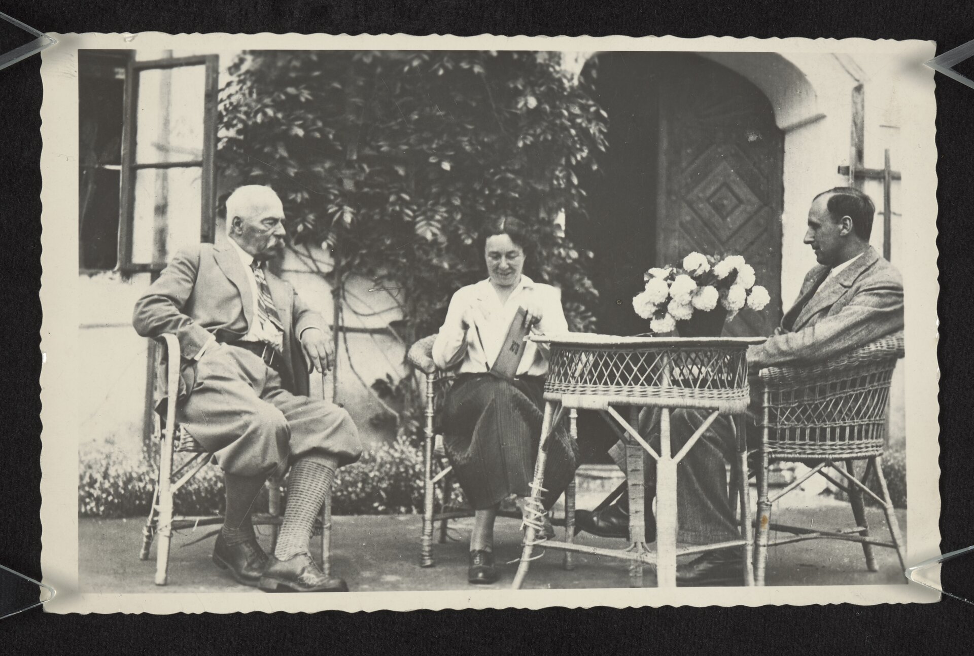 Czarnobiałe zdjęcie przedstawia troje ludzi siedzących na wiklinowych fotelach, przy wiklinowym stoliku przed domem. Po lewej stronie, starszy mężczyzna z siwym wąsem i siwymi wąsami. Ubrany w jasny garnitur, białą koszulę, na szyi ma chustkę w paski, spodnie ma podwinięte pod kolana, długie skarpety, czarne oksfordki. Po środku siedzi kobieta w średnim wieku, o ciemnych włosach, ubrana w białą koszulę, ciemną, długą spódnicę i oksfordki. Na kolanach trzyma książkę. Po prawej stronie, bokiem do kadru siedzi mężczyzna w średnim wieku. Ma ciemne, zaczesane na bok włosy, jasny garnitur z białą koszulą i ciemne buty. Na stoliku stoją białe kwiaty. W tle dom, widać drewniane drzwi z otwartym jednym skrzydłem, otwarte okno i rosnący na ścianie budynku bluszcz.