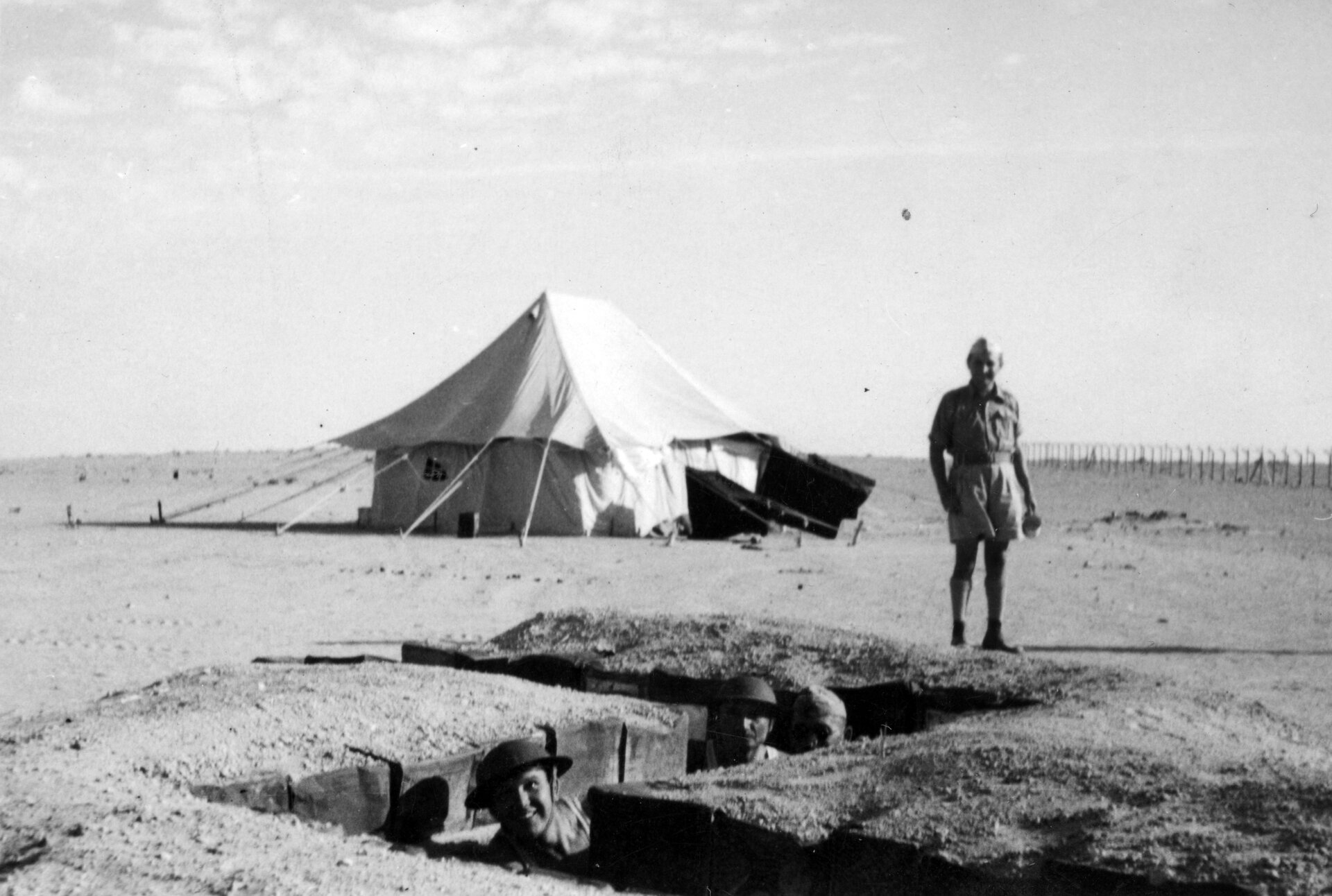 Zdjęcie przedstawia rozległą równinę. Na pierwszym planie znajduje się niewielki okop, w którym widoczne są głowy trzech polskich żołnierzy. Do okopu podchodzi czwarty żołnierz. W oddali znajduje się rozbity namiot.