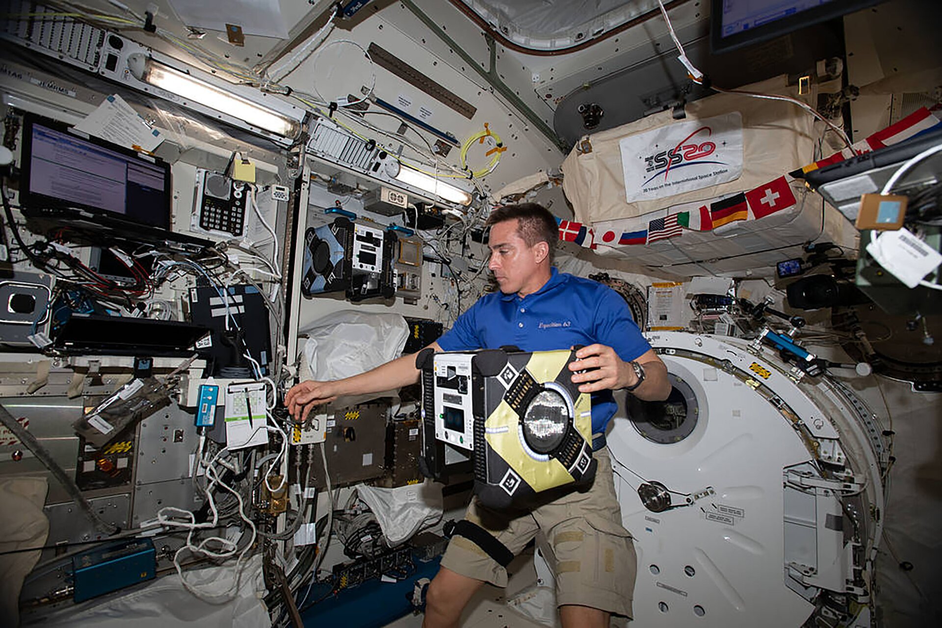 Rys. 2. Zdjęcie przedstawia astronautę w statku kosmicznym znajdującym się na orbicie okołoziemskiej. Astronauta unosi jedną ręką duże urządzenie.