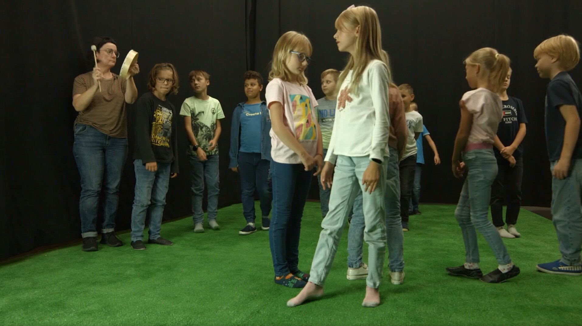 Fotografia przedstawia wnętrze sali o ciemnych ścianach i zielonej wykładzinie, na której stoi grupa dzieci oraz nauczycielka. Na pierwszym planie dwie blondwłose dziewczynki - obie ubrane w dżinsowe spodnie oraz jasne koszulki. Jedna spogląda się w podłogę, druga patrzy się w lewo. Na drugim planie nauczycielka grająca na bębenku za pomocą drewnianej pałeczki, obok niej stoją dzieci. Część spogląda się na kamerę umieszczoną za kadrem, a część spogląda się na nauczycielkę.