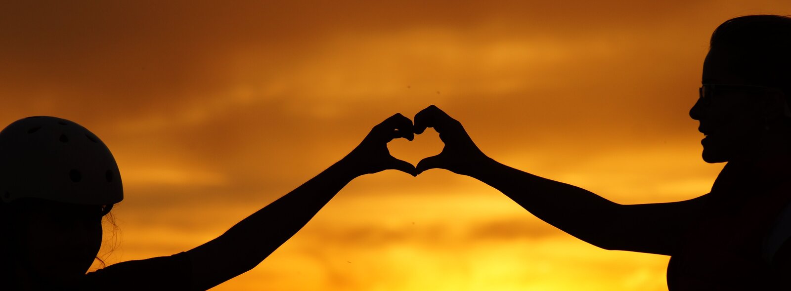 Zdjęcie przedstawia zaciemnione postacie kobiety i dziecka na tle żółto‑pomarańczowego nieba. Postacie wyciągają w swoim kierunku ręce. Ich dłonie tworzą symbol serca.