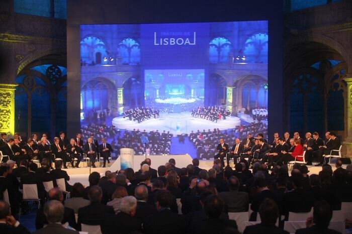 Zdjęcie przedstawia rzędy siedzeń ustawione półkoliście na scenie. Siedzą na nich ludzie. Na środku znajduje się ekran, na którym wyświetlana jest transmisja wydarzenia. Na górze ekranu jest napis Lisboa.