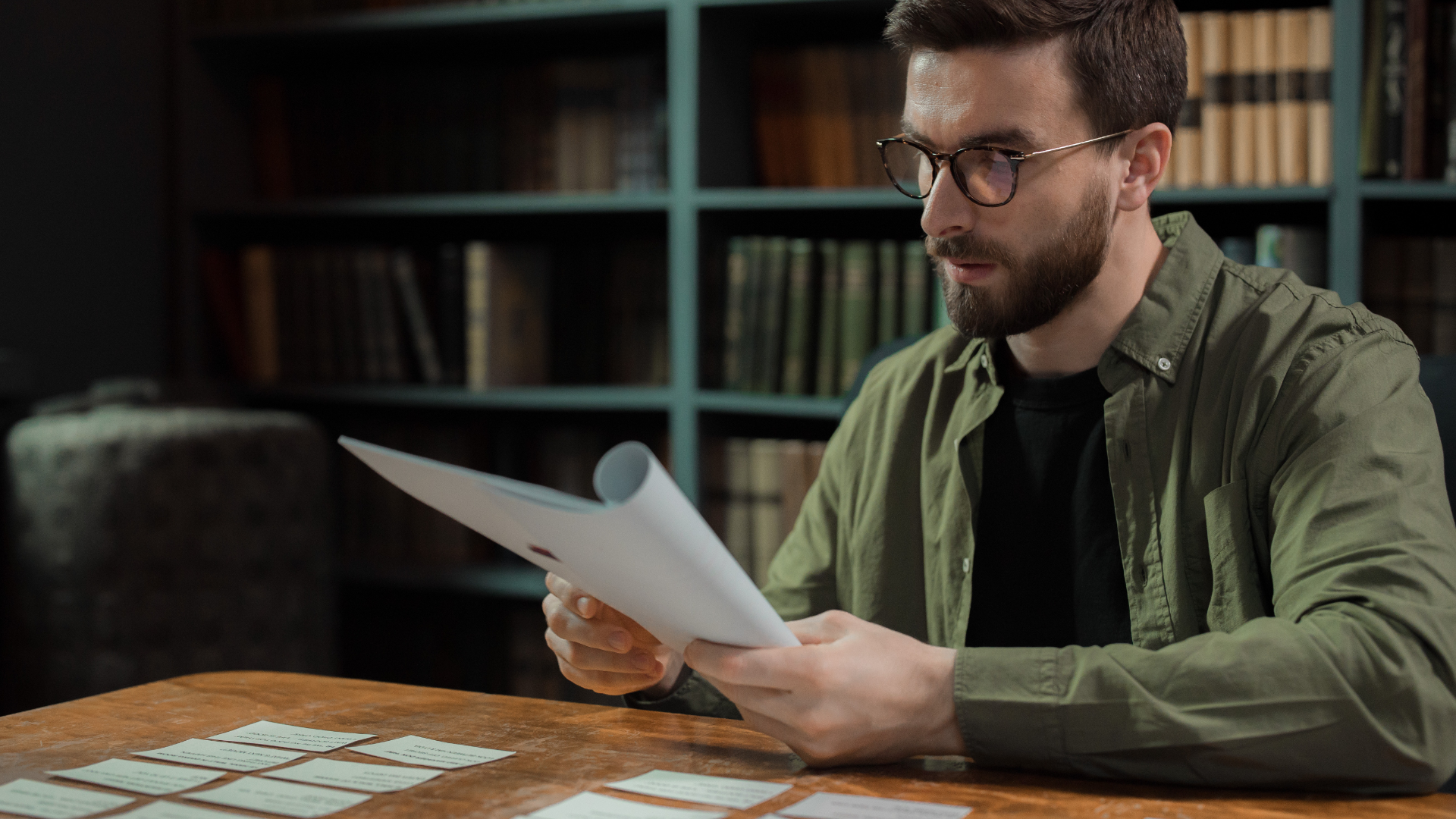 Zdjęcie przedstawia mężczyznę, który siedzi przy biurku. Mężczyzna ma na sobie rozpięta koszulę i okulary. W rękach trzyma dokument/scenariusz.