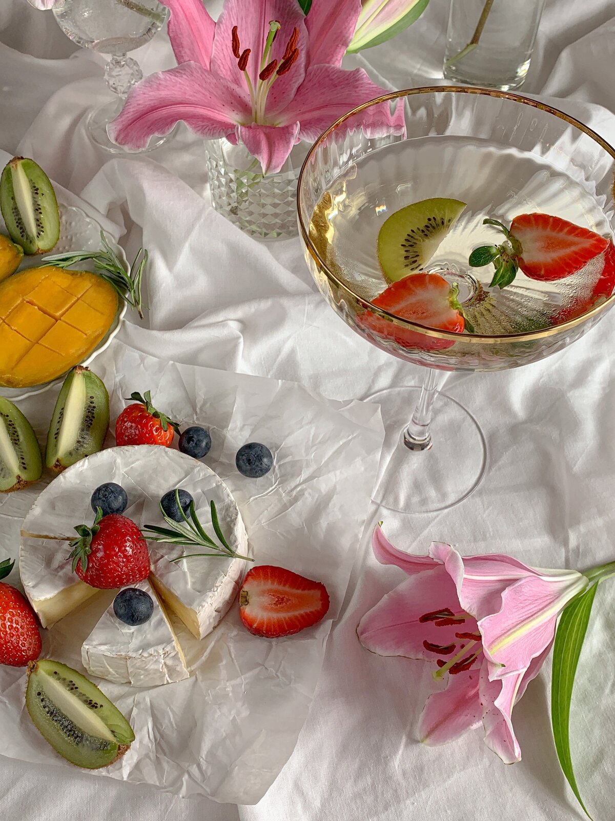 Na stole z lewej strony znajduje się pokrojony ser camembert, a wokół niego rozsypane truskawki, borówki, pokrojone cząstki owoców kiwi. Po prawej stronie szeroki kieliszek z białym winem, w którym pływają truskawki i kiwi. Obok kieliszka różowy kwiat.