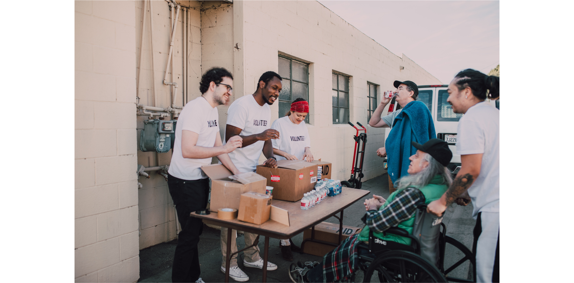 Zdjęcie przedstawia troje wolontariuszy, którzy rozdają butelki z wodą. Stoją przy stoliku. Na zdjęciu znajdują się jeszcze trzy osoby. Jeden z mężczyzn pije wodę z butelki, drugi siedzi na wózku inwalidzkim, a trzeci pcha ten wózek.