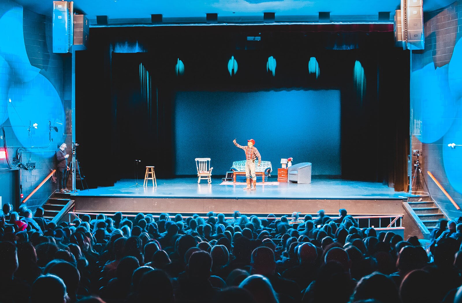 Zdjęcie przedstawia salę teatralną, z pełną widownią. Ujęcie zrobione z ostatnich rzędów siedzisk ukazuje głowy widzów od tyłu oraz scenę, na której stoi człowiek, a wokół niego krzesła i ławka.