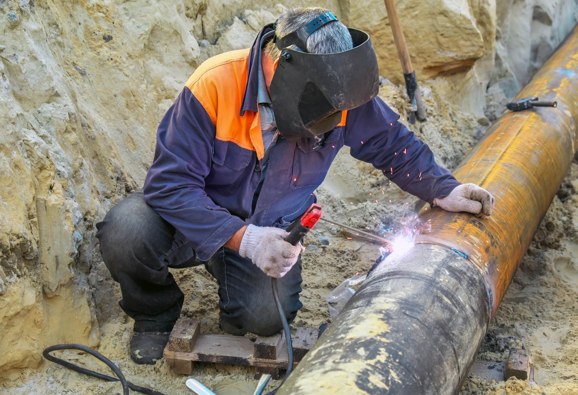Zdjęcie ukazuje mężczyznę w przyłbicy, który spawa stalową rurę umieszczoną w wykopie.