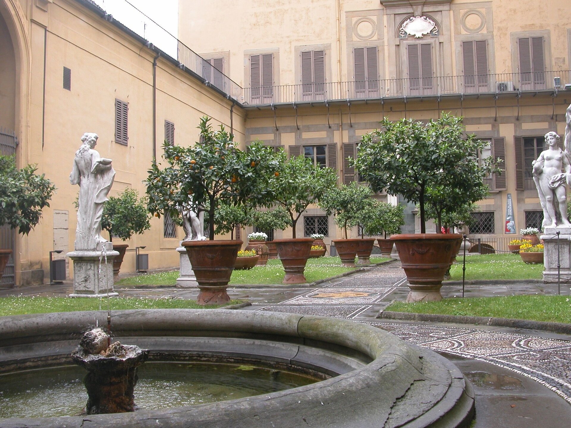Fotografia barwna przedstawiająca kwadratowy dziedziniec pałacu. Na środku znajduje się niewielka okrągła fontanna. Poza tym widoczne są rzeźby i rośliny.