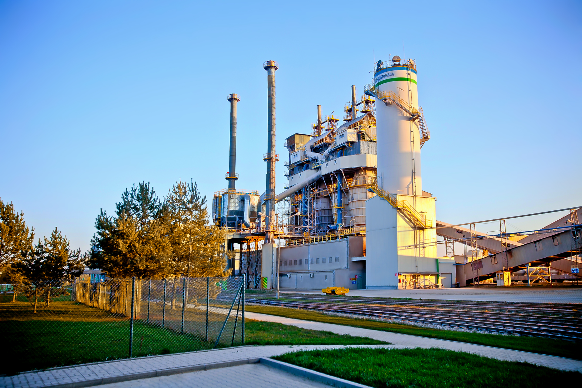 Zdjęcie zakładu wapienniczego, w którym wypala się wapień - Trzuskawica S.A. Na pierwszym planie widoczne tory, którymi przewożone są surowce i produkt. Za nimi stoi duży budynek fabryki z widocznymi kominami i instalacjami do produkcji i składowania wapna.