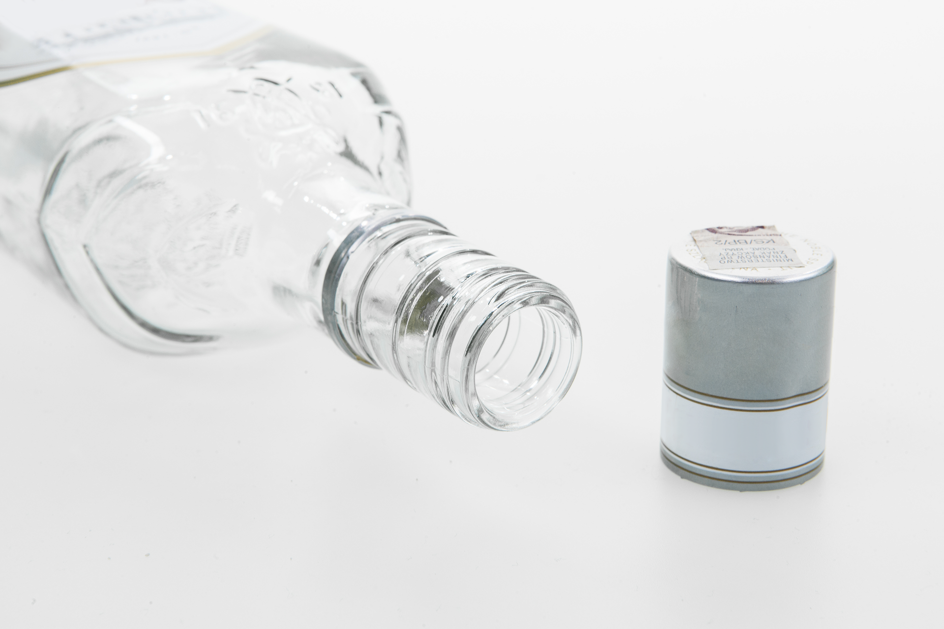 Drugie zdjęcie przedstawia leżącą szklaną butelkę skierowaną otworem w kierunku prawego rogu zdjęcia. Naprzeciw szara długa nakrętka butelki.