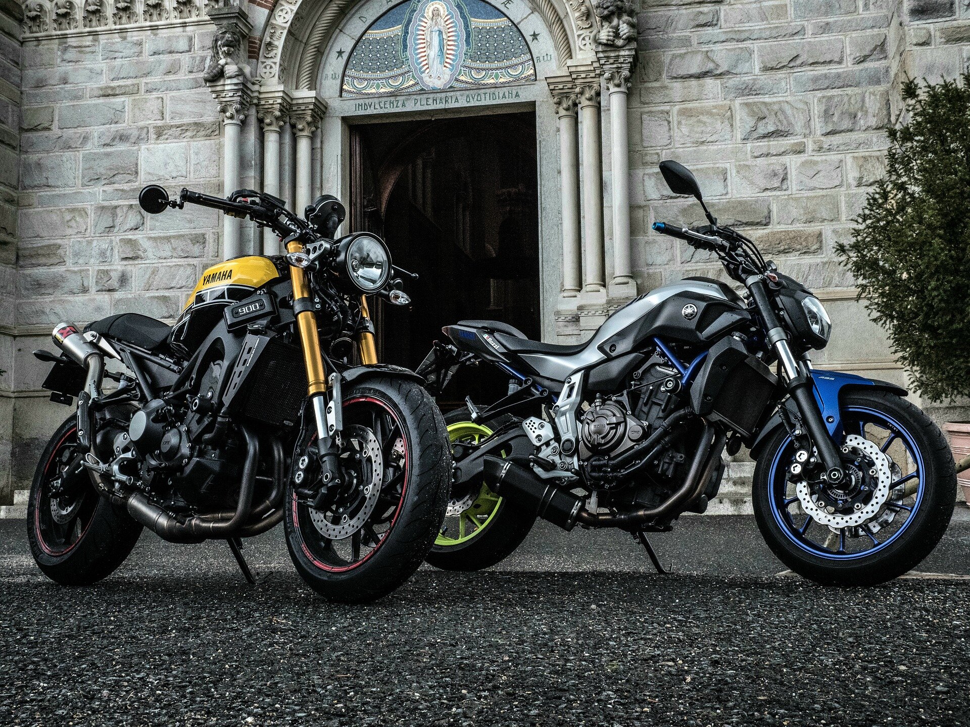 Zdjęcie przedstawia dwa zaparkowane przed kościołem motocykle. Jeden motocykl ma żółte dodatki, drugi niebieskie. Pojazdy są masywne. 