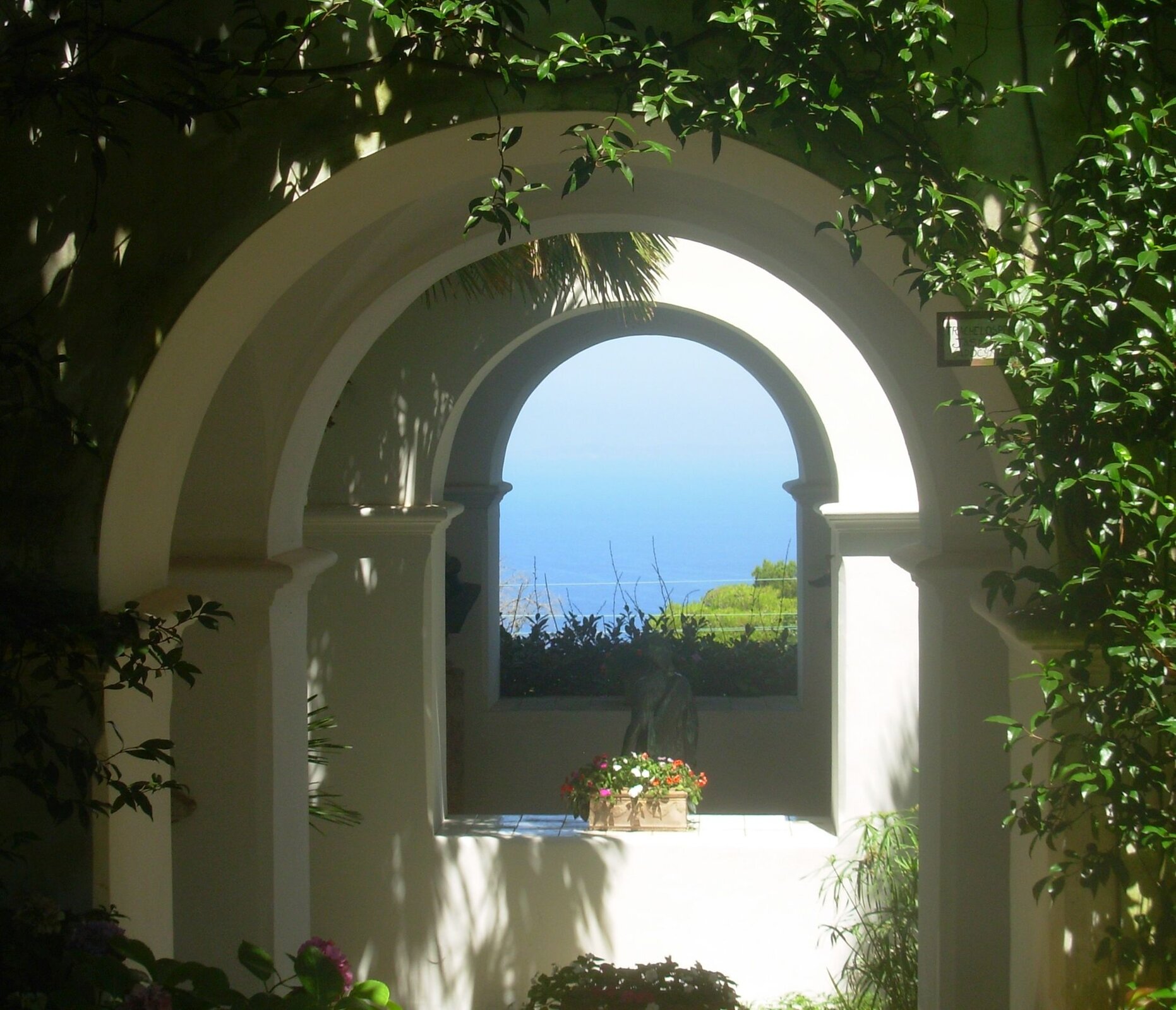 Fotografia autorstwa Elisabetty Monaco przedstawia widok na morze z Willi San Michele w Capri. W centrum fotografii znajdują się ceglane łuki oraz rzeźba mężczyzny i okno, z którego widoczne jest morze. Fotografia została wykonana w słoneczny dzień.