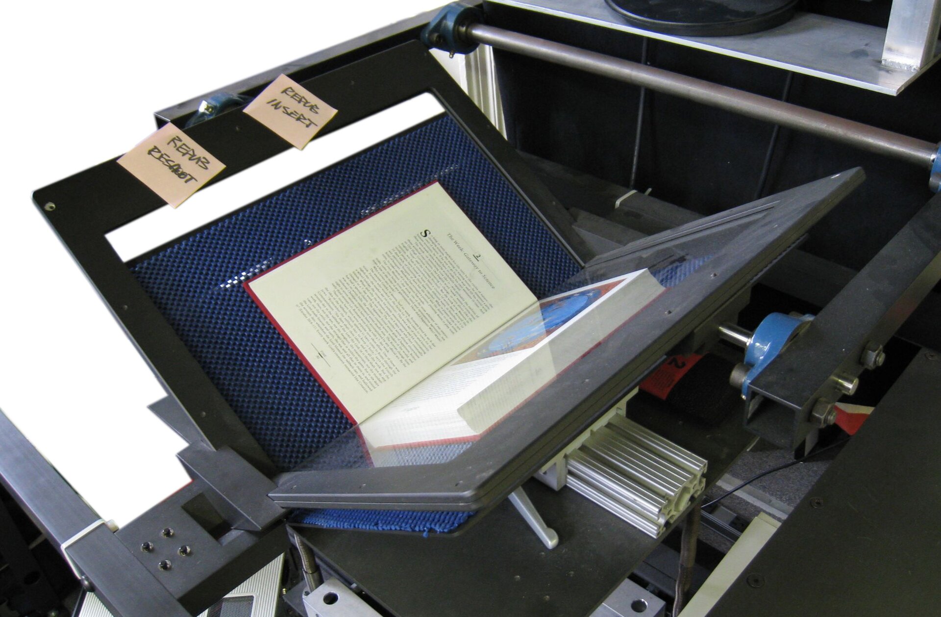 Zdjęcie przedstawia otwartą książkę ułożoną na dwuskrzydłowym skanerze do skanowania książek. Na lewym skrzydle skanera widoczne są poprzylepiane małe karteczki z napisami.