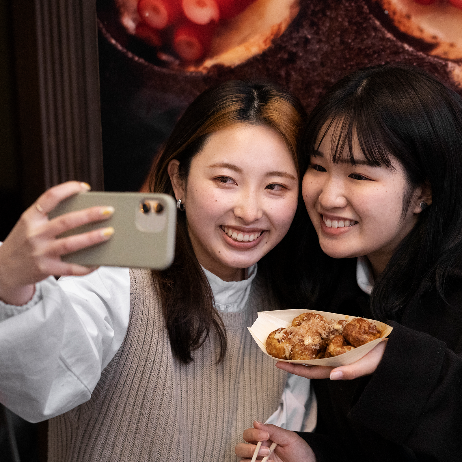 Zdjęcie przedstawia dwie młode, uśmiechnięte dziewczyny, w ujęciu od klatki piersiowej w górę. Dziewczyny stoją obok siebie i stykają się głowami. Dziewczyna po lewej stronie, z długimi, ciemnymi włosami, ubrana jest w białą bluzkę i beżowy pulower. W prawej wyciągniętej przed siebie ręce trzyma telefon komórkowy, którym robi zdjęcie. Dziewczyna po prawej stronie, z krótkimi, ciemnymi włosami, ubrana jest w czarną bluzkę i w lewej ręce - tuż przy brodzie – trzyma rożek wypełniony smażonym jedzeniem.