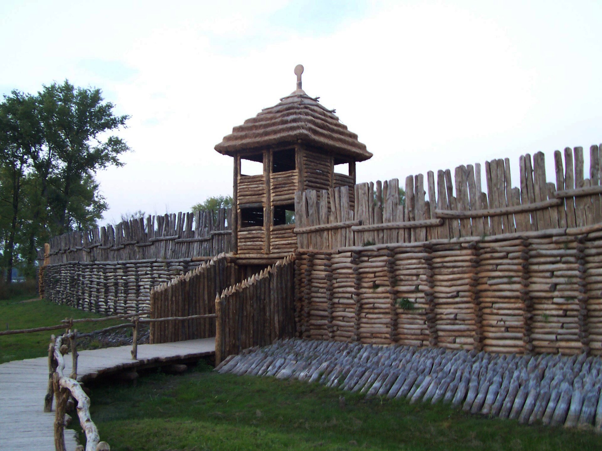 Zdjęcie przedstawia bramę i palisadę z drewna. Brama jest wysoko na kilkanaście metrów, góruje nad palisadą, która jest zbudowana z poziomych bali. Do bramy prowadzi kładka. 