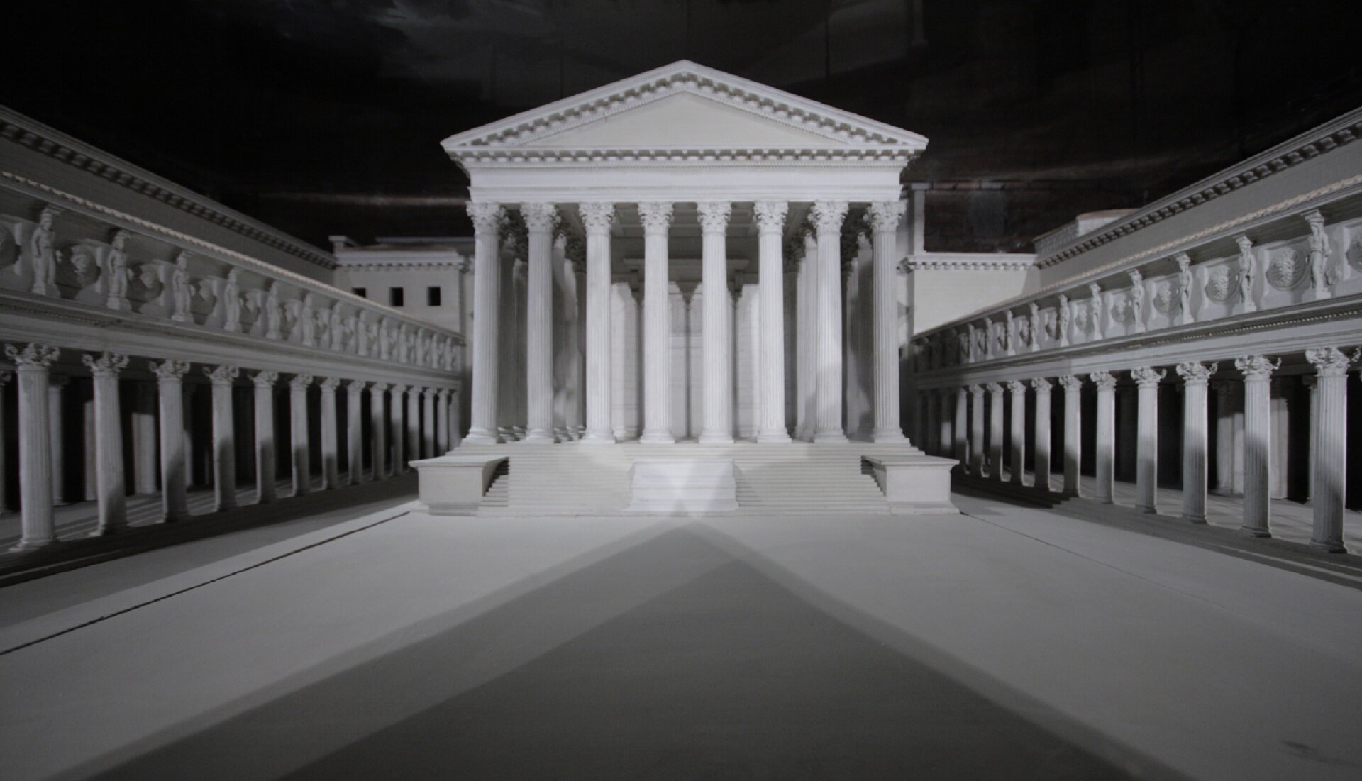 Ilustracja przedstawia makietę Forum Augusta. Na zdjęciu widać plac i znajdujący się na nim biały budynek w stylu rzymskim. Do budynku prowadzą schody. Przednią część stanowi portyk składający się z ośmiu kolumn zakończonych frontonem. Po bokach wzdłuż całego placu rozciągają się kolumny z zadaszeniem tworząc krużganek.