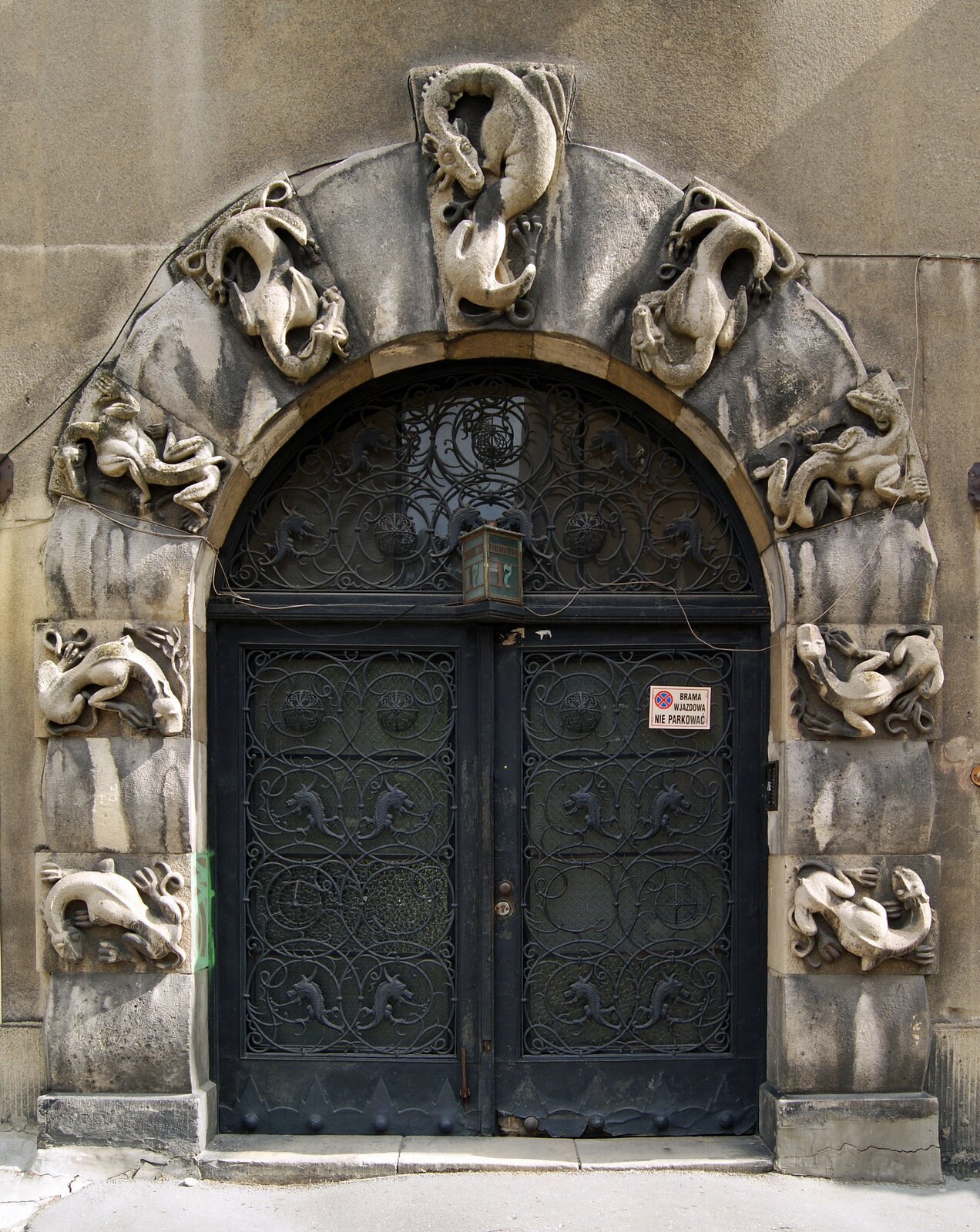 Zdjęcie przedstawia kamienny portal w kształcie łuku, który składa się z prostokątnych bloków. Na co drugim bloku umieszczono rzeźbę stworzenia przypominającego smoka. Portal okala szklane drzwi okute ażurową metalową dekoracją.