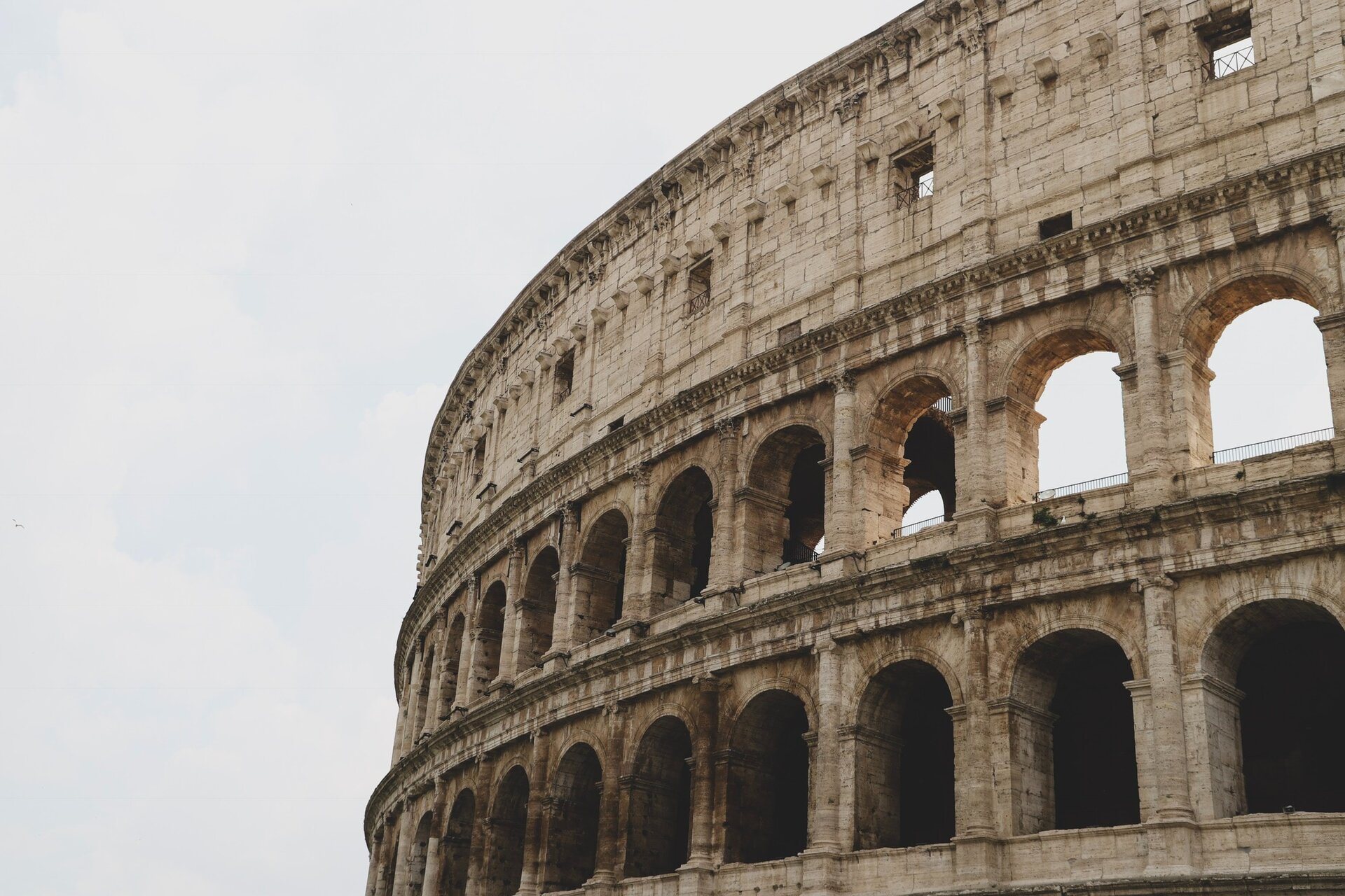 Zdjęcie przedstawia przykładową atrakcję turystyczną, w tym przypadku fragment ruin Koloseum w Rzymie. Budowla widoczna jest po prawej stronie zdjęcia. Całość na tle szarego nieba.