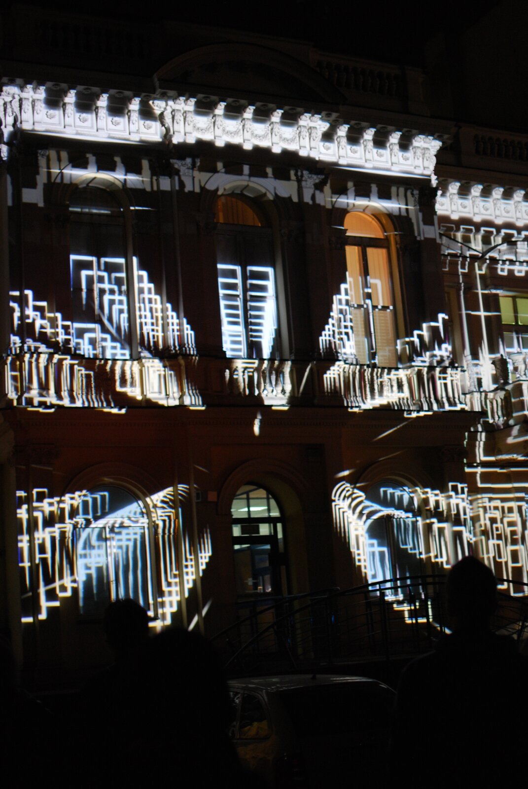 Ilustracja przedstawia prezentację 3D mappingu w Łodzi. Ukazuje wyświetloną na fasadzie budynku przestrzenną kompozycję, złożoną z ustawionych jedna za drugą linearnych konstrukcji, które tworzą perspektywiczny efekt stojących świetlnych ram. 