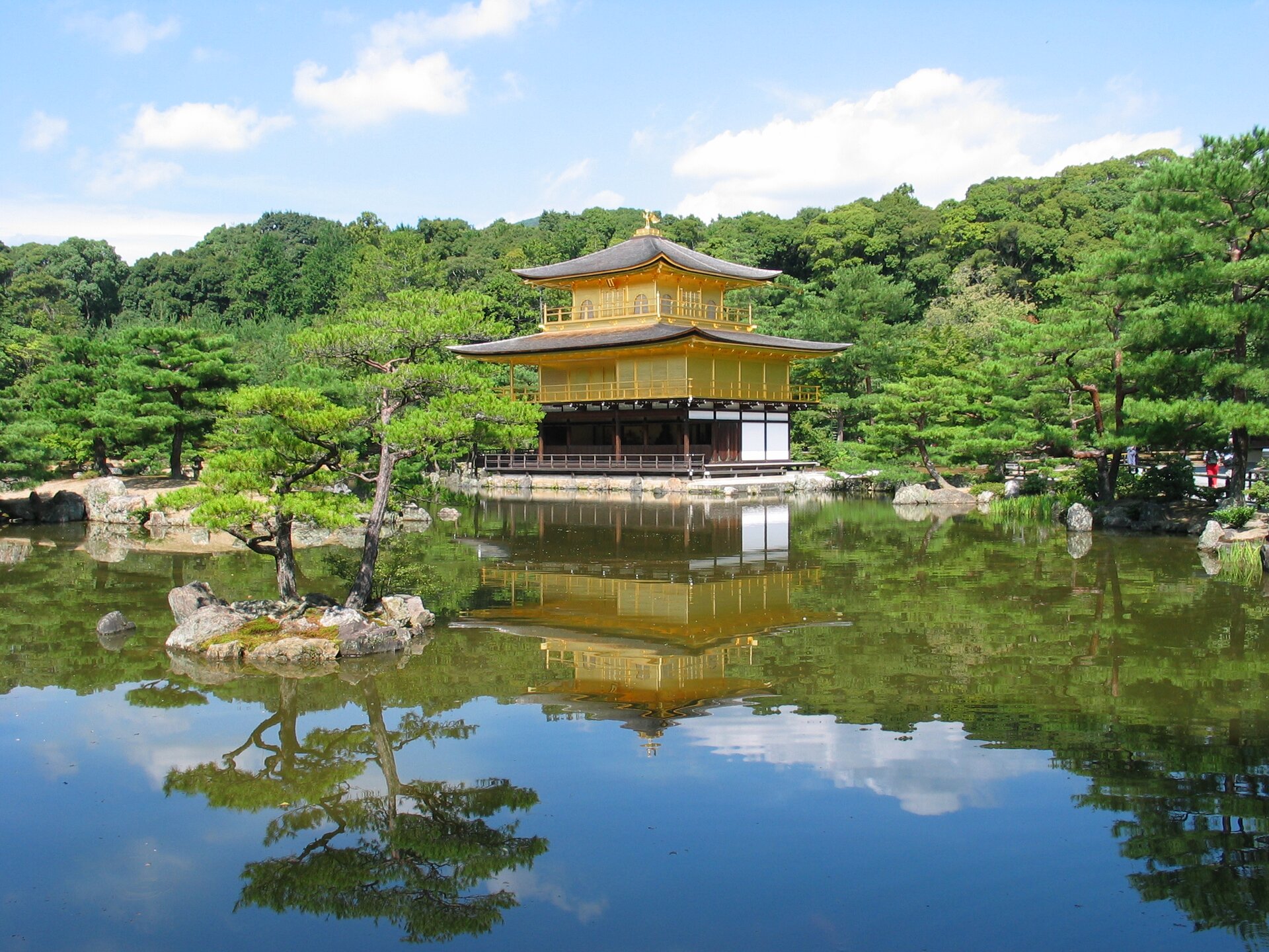 Na zdjęciu typowa japońska świątynia udekorowana złotem. Położona nad wodą w parku. Bujna roślinność.