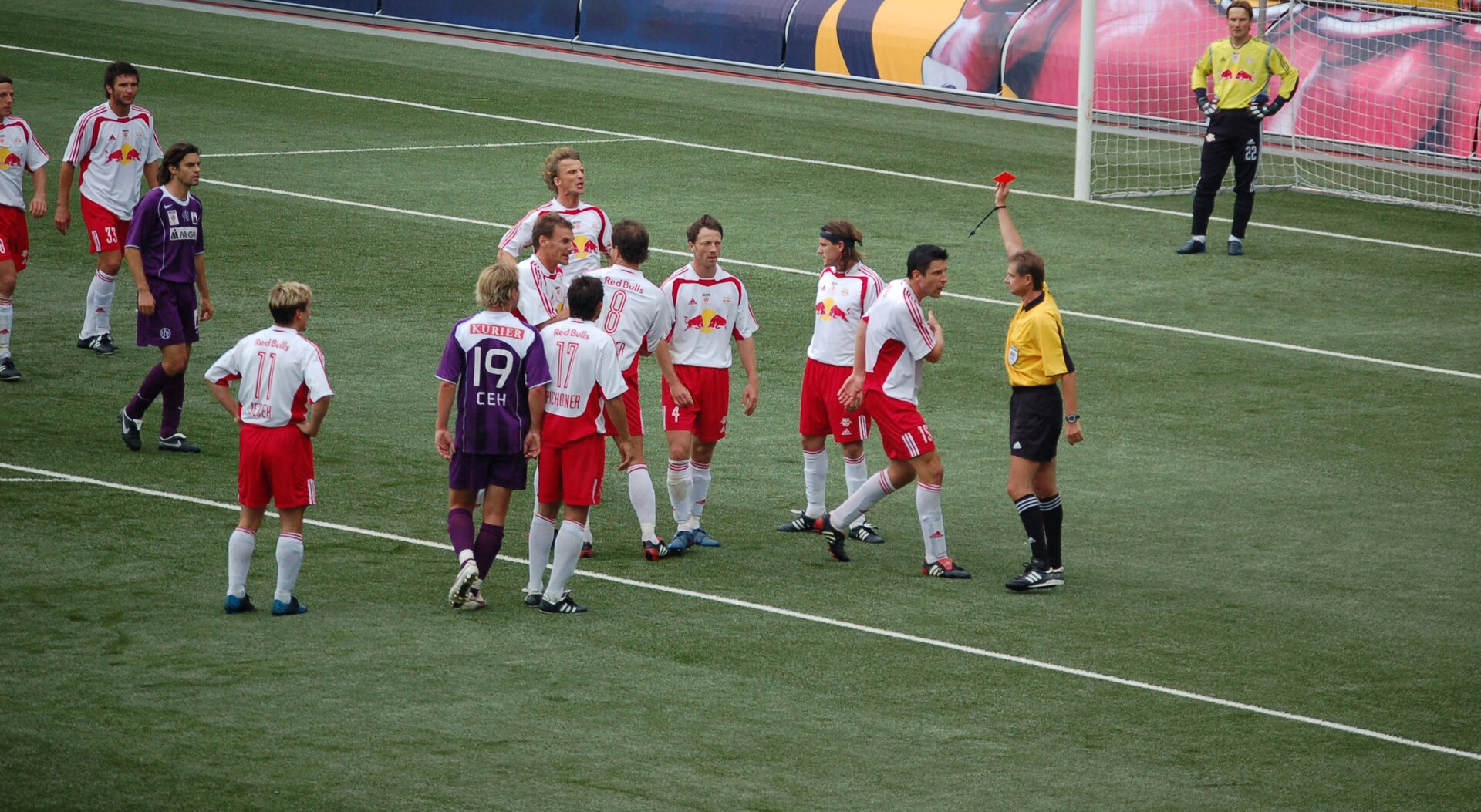Ilustracja przedstawia boisko piłkarskie z widownią pełną kibiców. Na boisku na polu karnym stoją zawodnicy obu drużyn. Scena ukazuje moment, w którym jeden z zawodników otrzymuje czerwoną kartkę od sędziego.