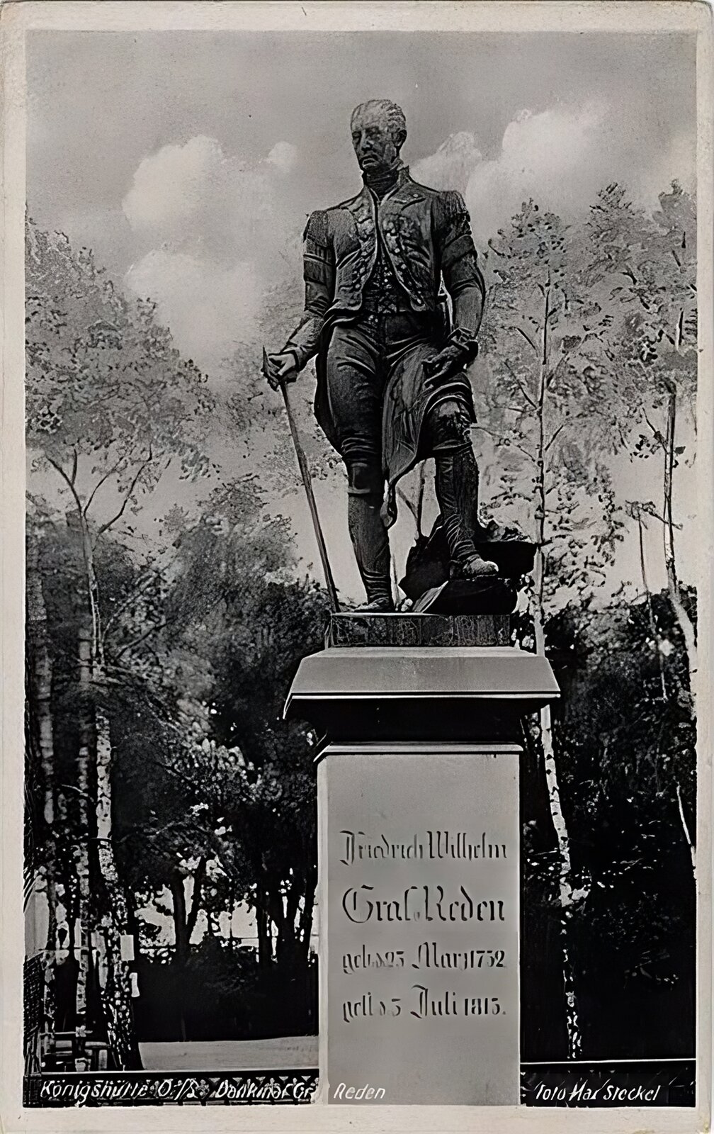 Kliknij, aby powiększyć Zdjęcie przedstawia pomnik z postacią hrabiego Friedricha Wilhelma von Redena w mundurze górniczym, który prawą ręką opiera na kolanie, na którym leży rozwinięta mapa, a prawą opiera na górniczym oskardzie. Pod prawą jego stopą leży bryłka rudy. Posąg z brązu ustawiony jest na cokole z jasnoszarego marmuru. Na przodzie cokołu jest inskrypcja w języku niemieckim, która brzmi: Fryderyk Wilhelm hrabia von Redenur. 23 marca 1752, zm. 3 lipca 1815. Na tylnej ścianie cokołu jest inskrypcja w języku niemieckim, która brzmi: Założycielowi śląskiego górnictwa wdzięczny przemysł górniczy i hutniczy oraz robotnicy Śląska 1852. W tle znajdują się wysokie drzewa (brzozy).