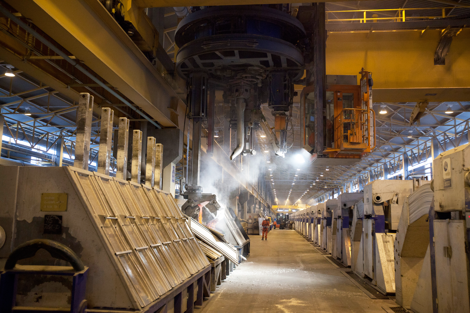 Zdjęcie elektrolizera, wielkiego urządzenia fabrycznego, w którym prowadzona jest elektroliza tlenku glinu na skalę przemysłową. W centrum ilustracji znajduje się szerokie przejście, a z obu jego stron poustawiane są metalowe zbiorniki. Powyżej umieszczone są betonowe balkony z żółtymi barierkami.