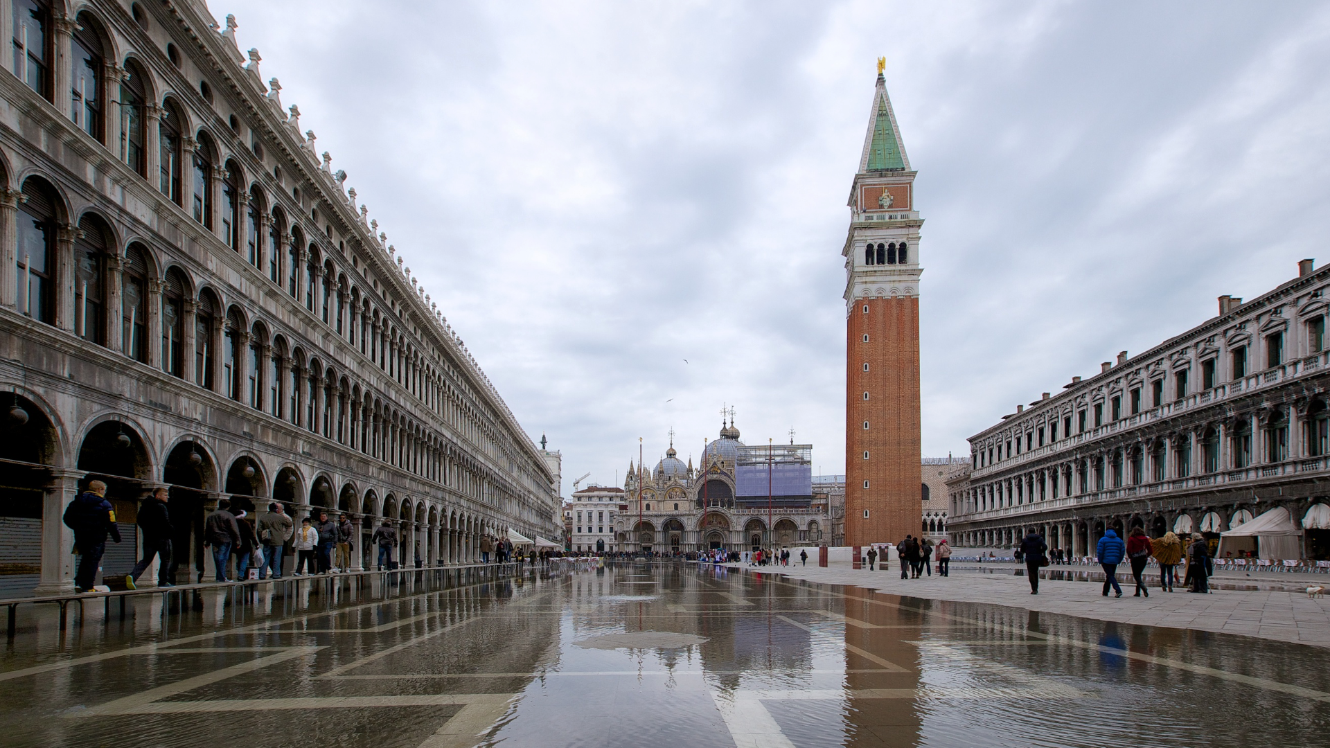 Zdjęcie przedstawia zalany wodą Plac Świętego Marka w Wenecji. Wokoło placu widoczni są spacerujący ludzie.