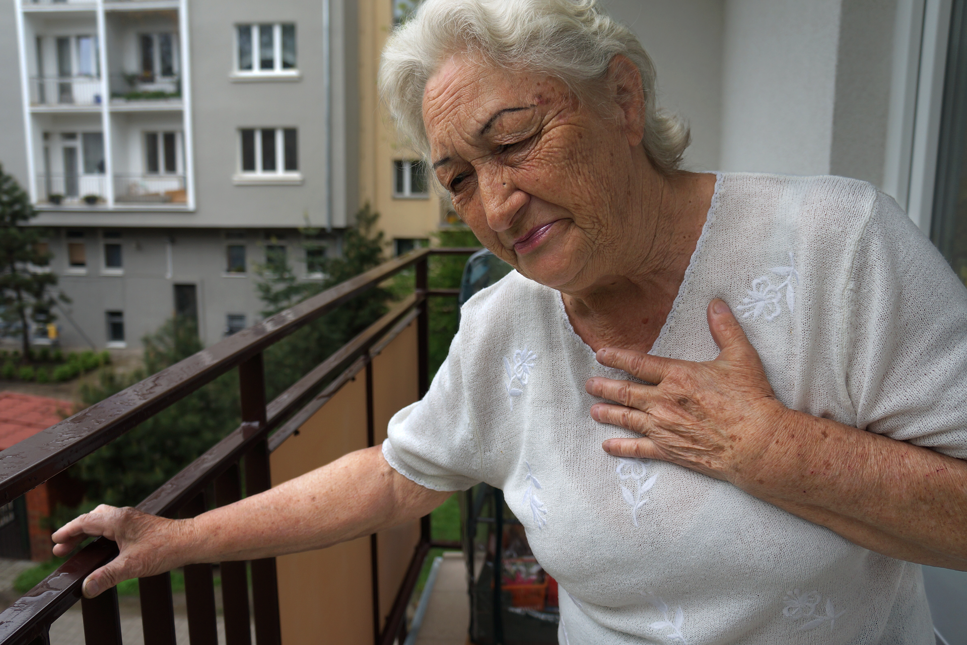 Kolorowe zdjęcie przedstawia starszą kobietę stojącą na balkonie. Kobieta jest zwrócona przodem do obserwatora, prawą ręką wspiera się o metalową balustradę balkonu, a lewą dłoń przycisnęła do prawej strony klatki piersiowej. Ma przymknięte oczy. Twarz kobiety pokryta jest licznymi zmarszczkami. Jej usta są wąskie, zaciśnięte od bólu. Kobieta pochyla się lekko w lewo – w stronę balustrady.