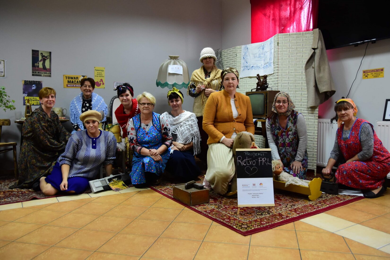 Zdjęcie przedstawia warsztaty artystyczne w Gminnym Ośrodku Kultury w Gostycynie. Na ilustracji znajduje się grupa kobiet siedzących na dywanie. Niektóre z kobiet ubrane są w fartuchy. Przed kobietami znajduje się tabliczka z nazwą projektu Retro- PRL. W tle ilustracji znajduje się pokój, na którego ścianie wiszą różne plakaty. 
