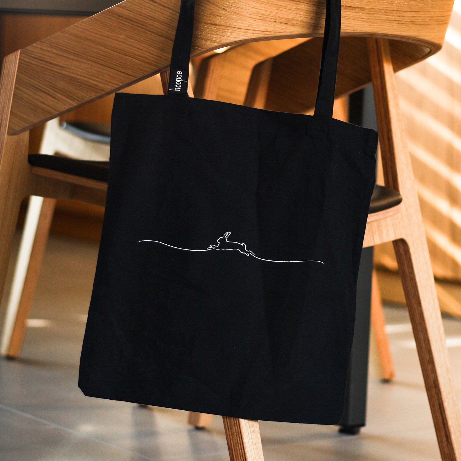  Zdjęcie przedstawia czarną materiałową torbę wiszącą na oparciu drewnianego krzesła. Pośrodku torby widoczny jest biały kontury przedstawiający skaczącego królika.