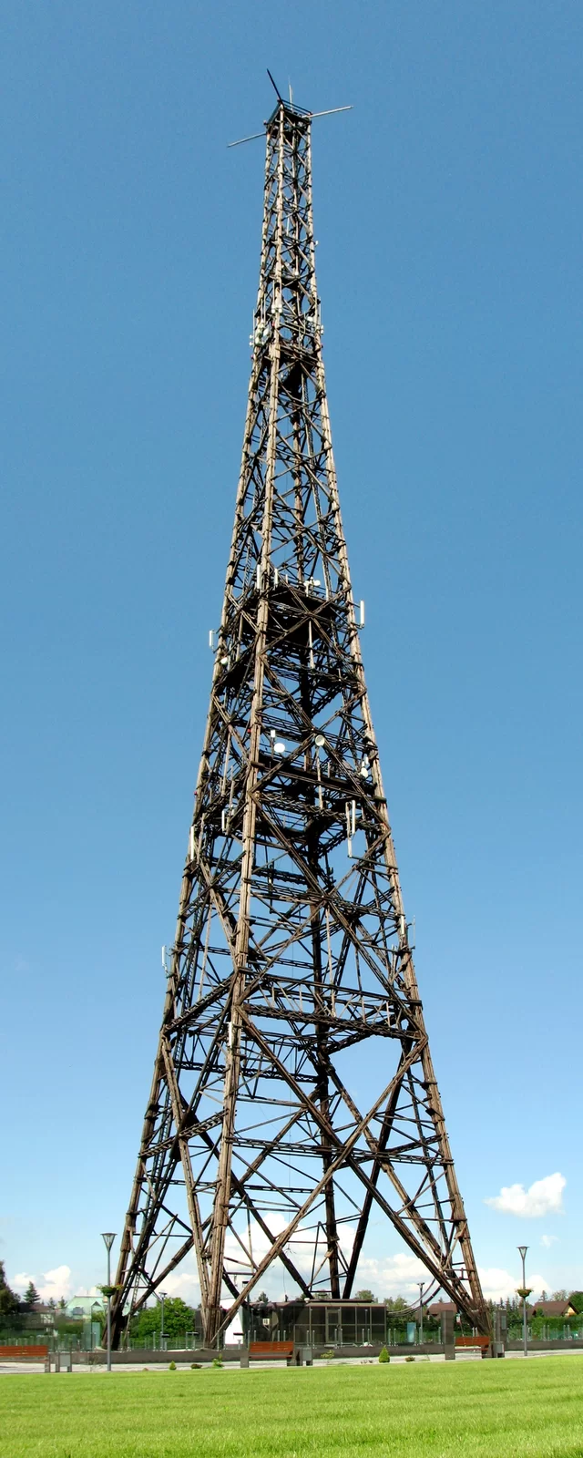 Zdjęcie przedstawia wieżę radiostacji. Konstrukcja drewniana. Wysoka wieża widziana z perspektywy człowieka stojącego na ziemi. Na dole widać trawnik, na górze szczyt wieży na tle nieba.