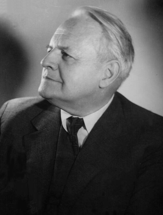 Zdjęcie przedstawia lewy profil starszego mężczyzny w garniturze. Mężczyzna ma pełną twarz, wysokie czoło. Jego włosy są proste, siwe, zaczesane do tyłu. Mężczyzna ubrany jest w garnitur, koszulę i krawat.