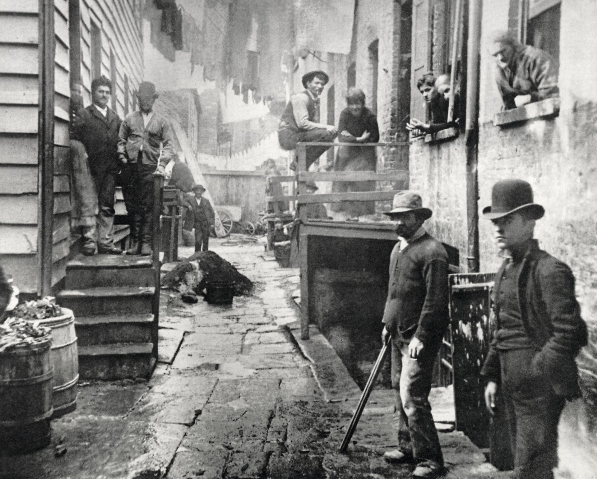 Zdjęcie przedstawia slumsy na Manhattanie. W ciasnej uliczce stoją mężczyźni w kapeluszach. Po obu stronach uliczki znajdują się podesty ze schodami. Również na nich stoją mężczyźni. W budynku z prawej strony z okien wychylają się kolejne osoby.