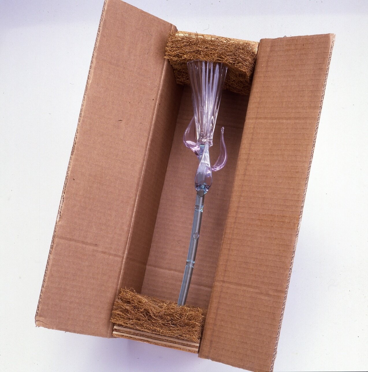 Zdjęcie przedstawia ozdobne, szklane naczynie w opakowaniu z kartonu. W górnej i dolnej części zabezpieczone jest kształtkami z włókna kokosowego.