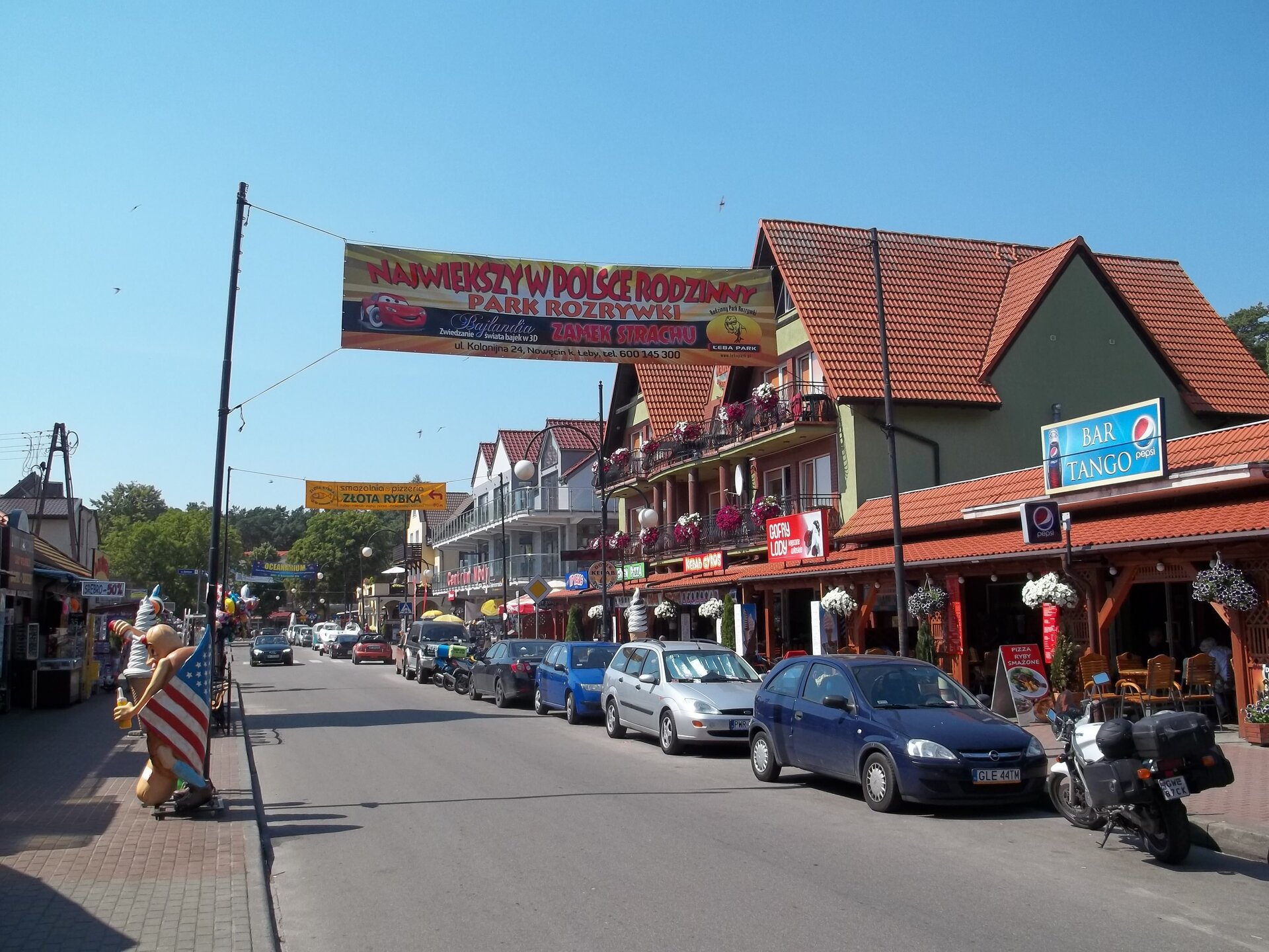 Zdjęcie przedstawia ulicę, na której są zaparkowane samochody. Po lewej i prawej stronie drogi umieszczone są bary czy budki z lodami, natomiast za nimi znajdują się osobne budynki mieszkalne. Na ulicy w dalszej części ilustracji ukazane jest auto. Przy drodze zamieszczone są reklamy różnych atrakcji turystycznych. 