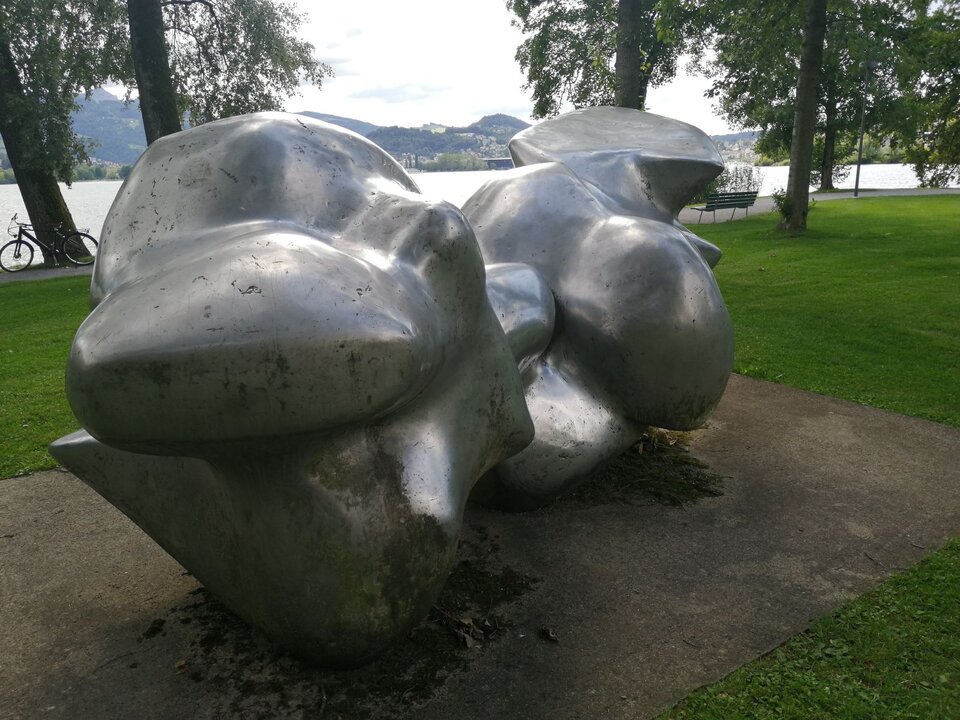 Zdjęcie przedstawia rzeźbę współczesną wykonaną z polerowanego metalu. Usytuowana jest w poziomie, w miejscu publicznym wśród zieleni. Jego abstrakcyjna forma złożona jest z wielu złączonych ze sobą, obłych kształtów pokaźnych rozmiarów.   