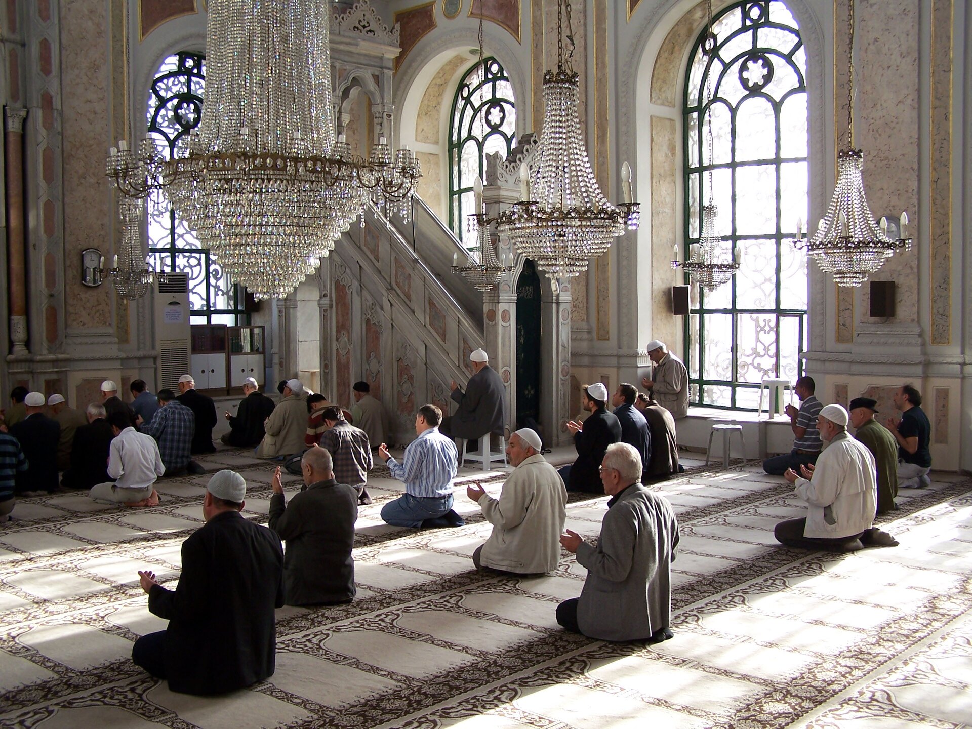 Zdjęcie przedstawia wnętrze meczetu. Na podłodze siedzą sami mężczyźni. Zwróceni są w jedną stronę. Mają lekko uniesione na boki ręce w geście  modlitwy. Nad modlącymi się wiszą duże, kryształowe żyrandole. 