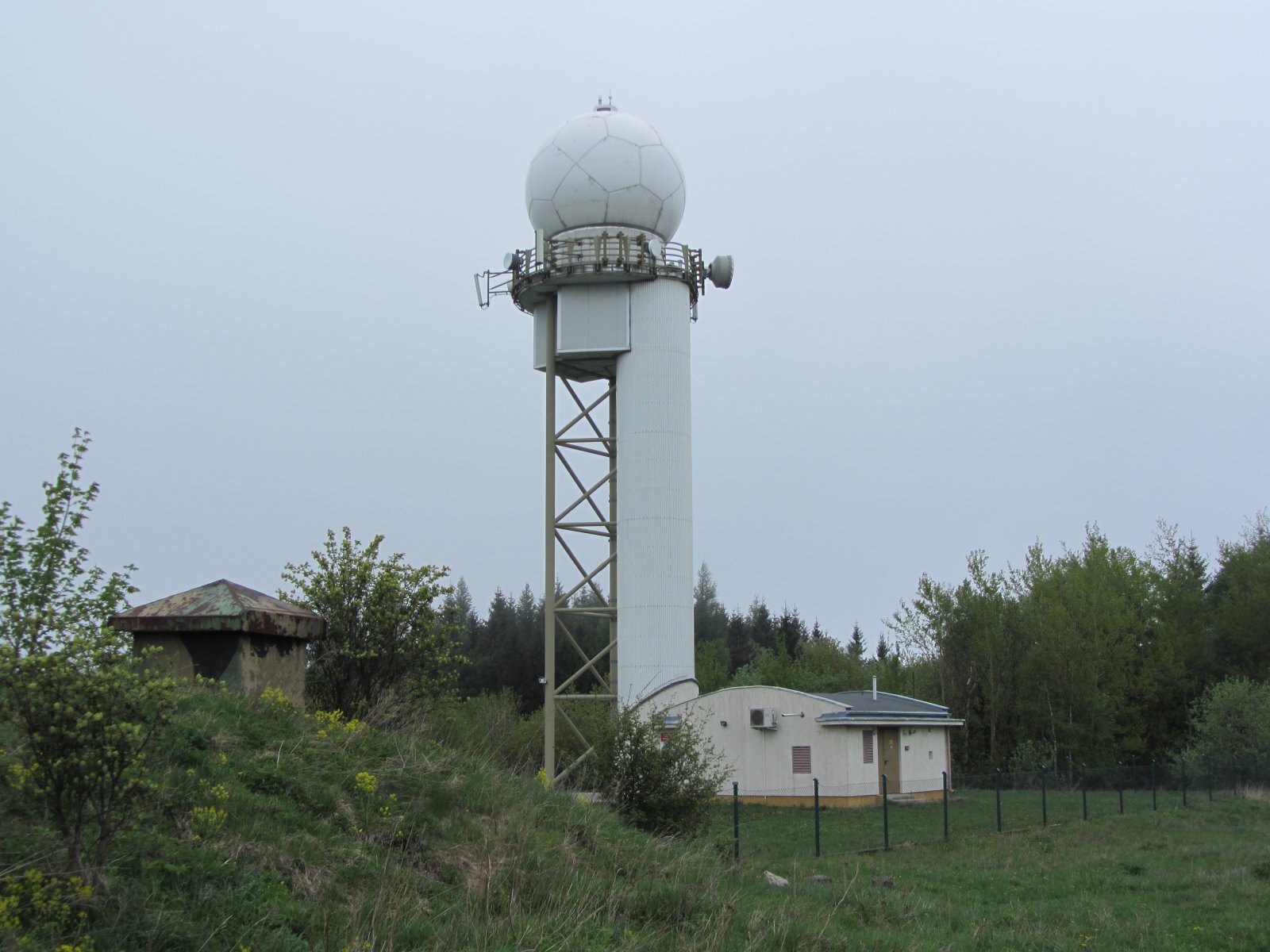 Zdjęcie przedstawia radar w Pastewniku. Ma kształt kuli. Znajduje się wysoko nad ziemią. Do konstrukcji przylega budynek.