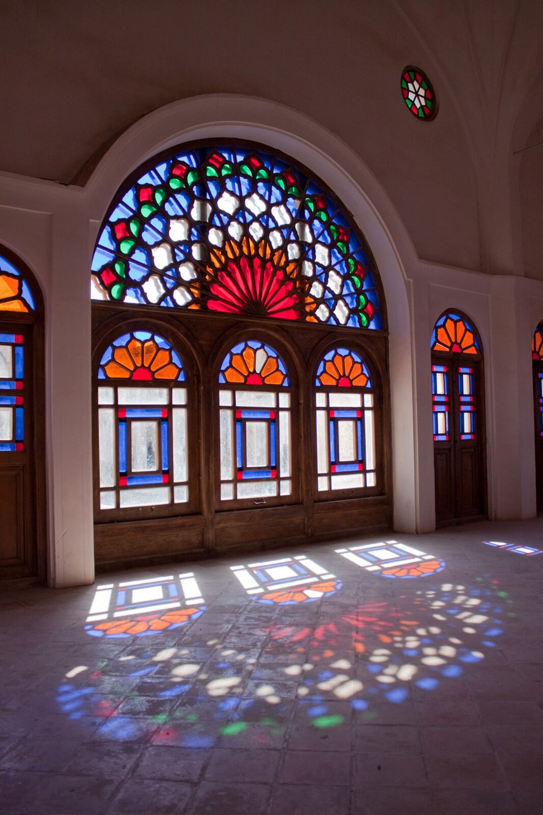 Zdjęcie przedstawia ścianę wejściową do kościoła z głównym i dwoma bocznymi wejściami, ozdobionymi witrażami. Wpadające przez nie światło odbija się w posadzce, tworząc barwne odbicie wejścia.