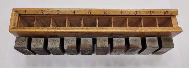 Na zdjęciu widoczna jest skala wzorców Jernkontoret. W drewnianym pudełku widocznych jest dziesięć otworów. Nad otworami znajdują się cyfry od jeden do dziesięć. Przed pudełkiem wyciągnięte są prostopadłościenne wzorce stali. Na górnej powierzchni wzorca próbki widoczne są różnice w wielkości ziarna. Próbka oznaczona cyfra jeden ma najgrubsze ziarno a próbka oznaczona cyfrą dwa najmniejsze.