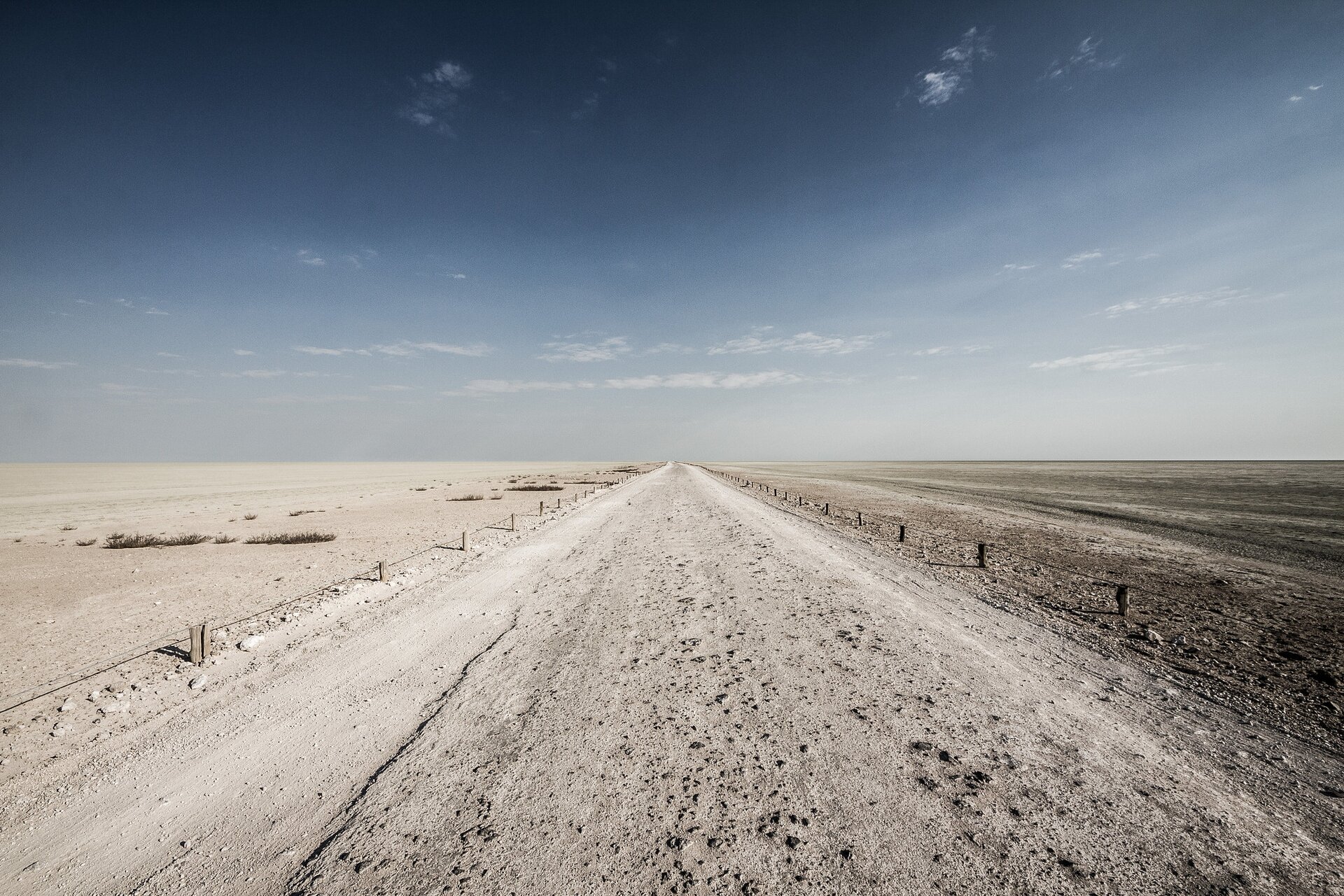 Na zdjęciu szeroka, nieutwardzona droga. Ma jasnoszary kolor, podobnie jak płaski teren po bokach. Jałowy obszar, niemal brak roślinności.