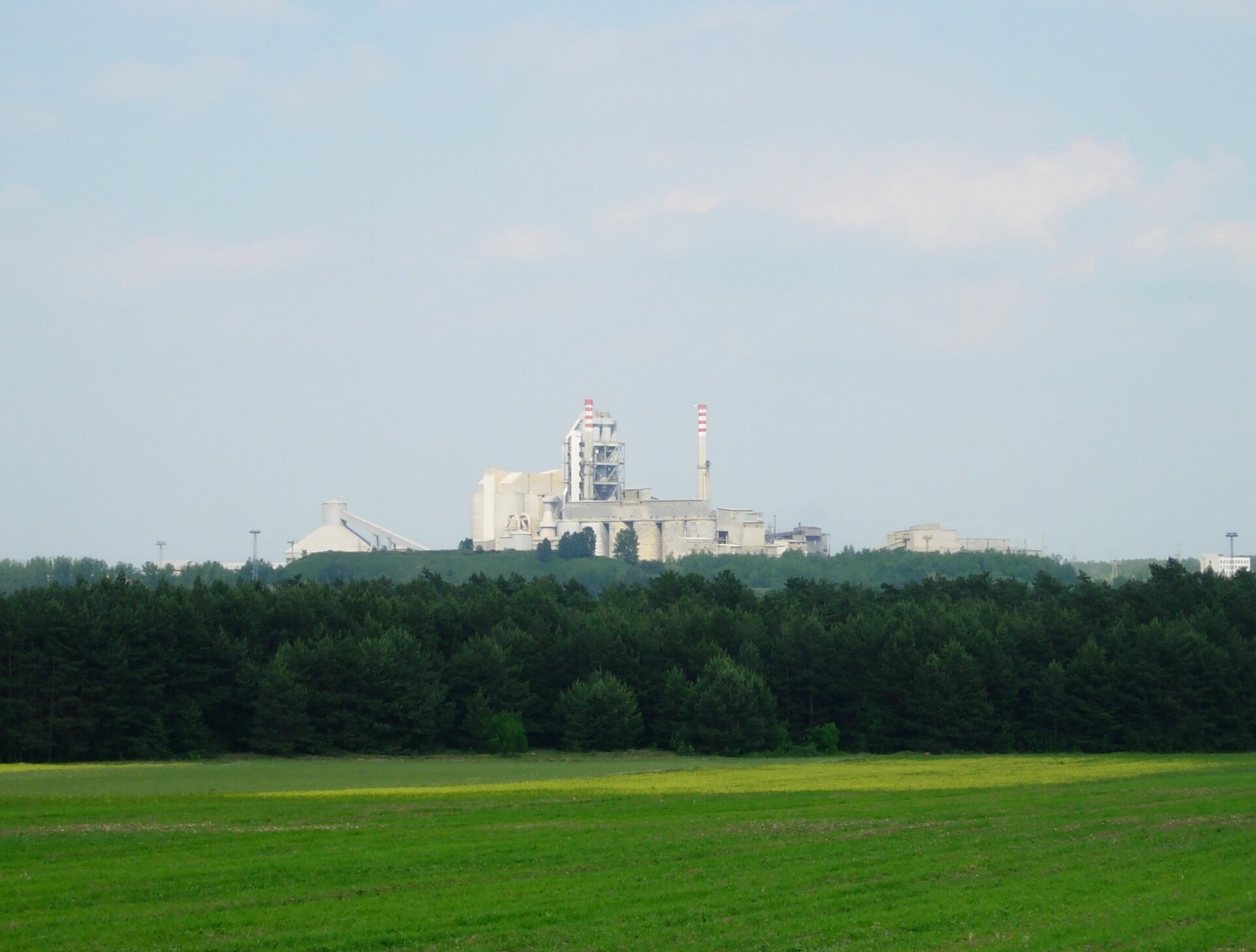 Na zdjęciu zabudowania przemysłowe. Na pierwszym planie pole i las.